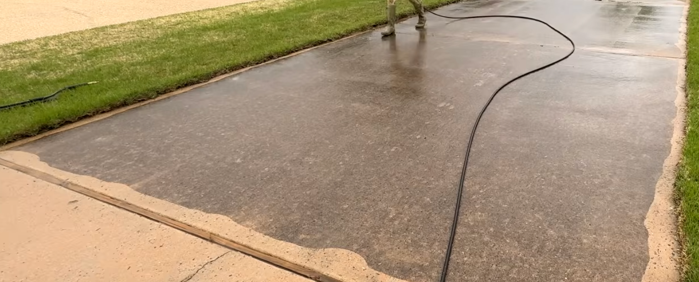 A person power washing a concrete walkway next to a grassy area. The walkway is wet and dark, the grass is green.