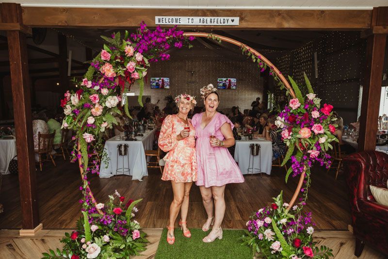 Two women are standing next to each other in front of a floral arch.