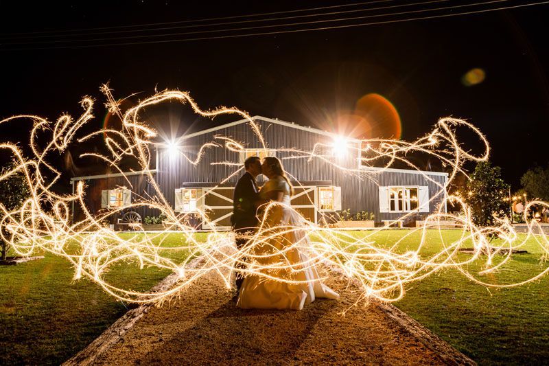 A bride and groom are kissing in front of Aberfeldy Barn at night surrounded by sparklers.
