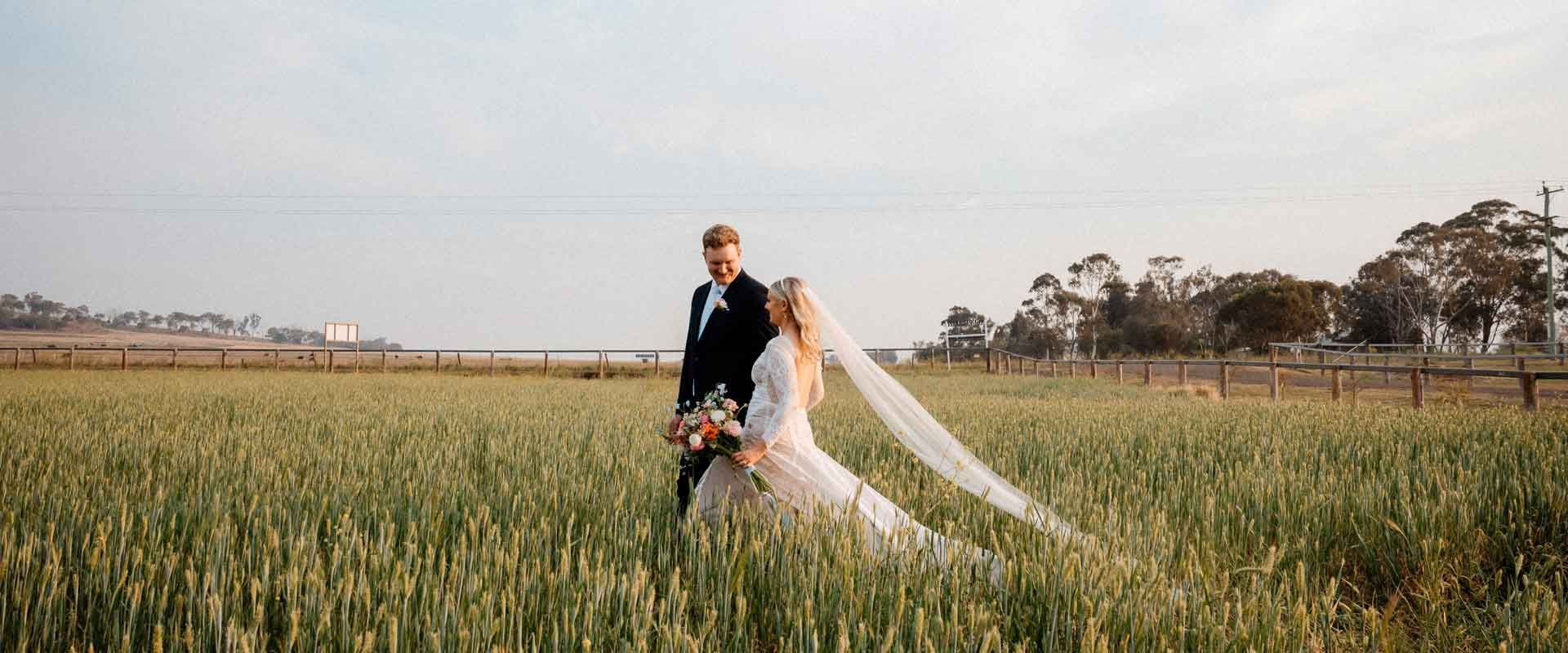 A newlywed couple walks through a field at Aberfeldy Barn; the bride wears a long veil.