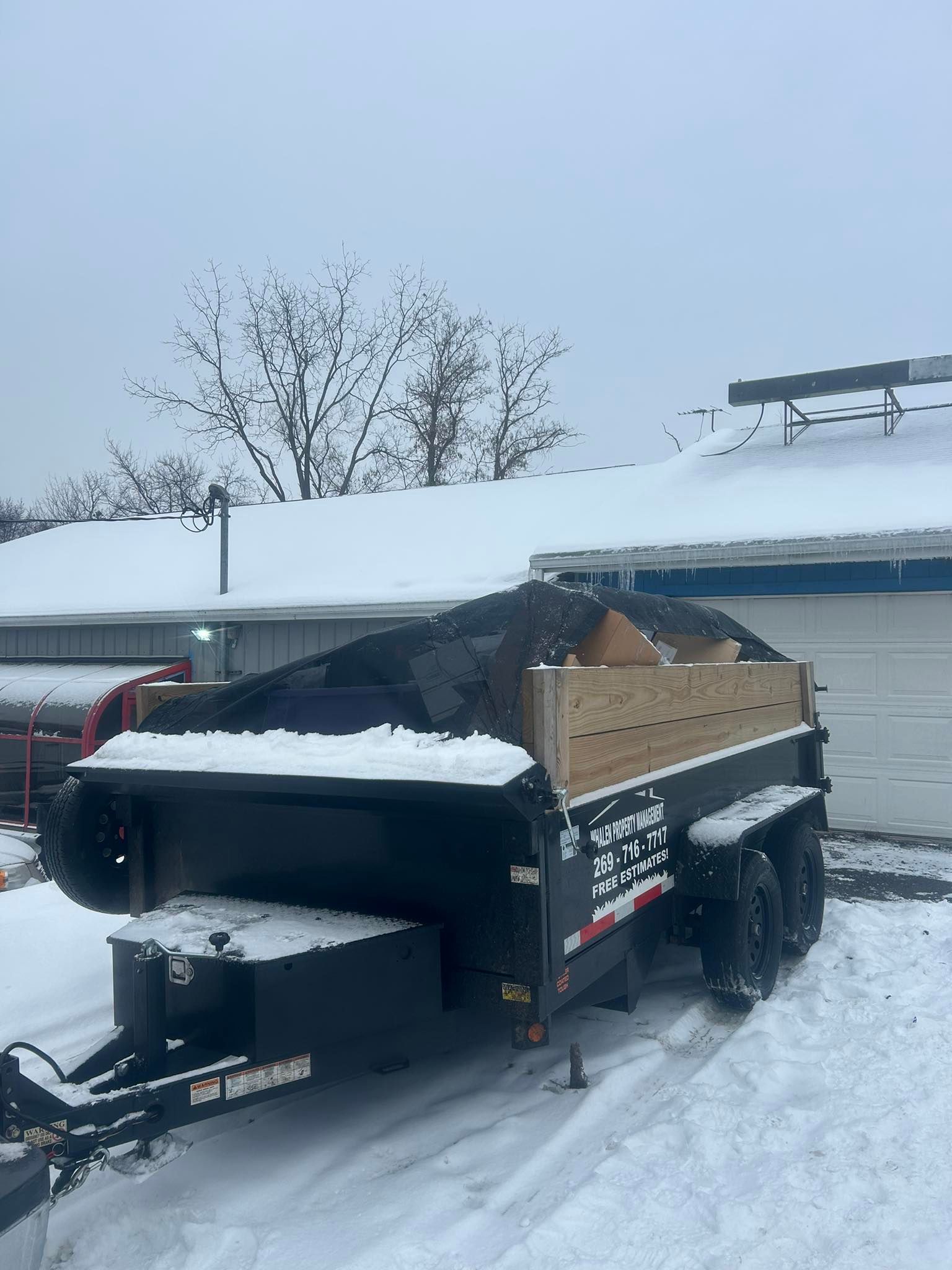 A black dump trailer with wooden side extensions, loaded with debris, parked in a snow-covered driveway.