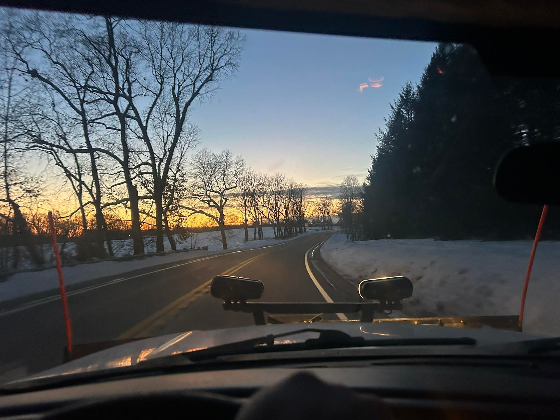 View from the driver's seat of a snowplow during a sunset, with a snowy landscape lining the road.