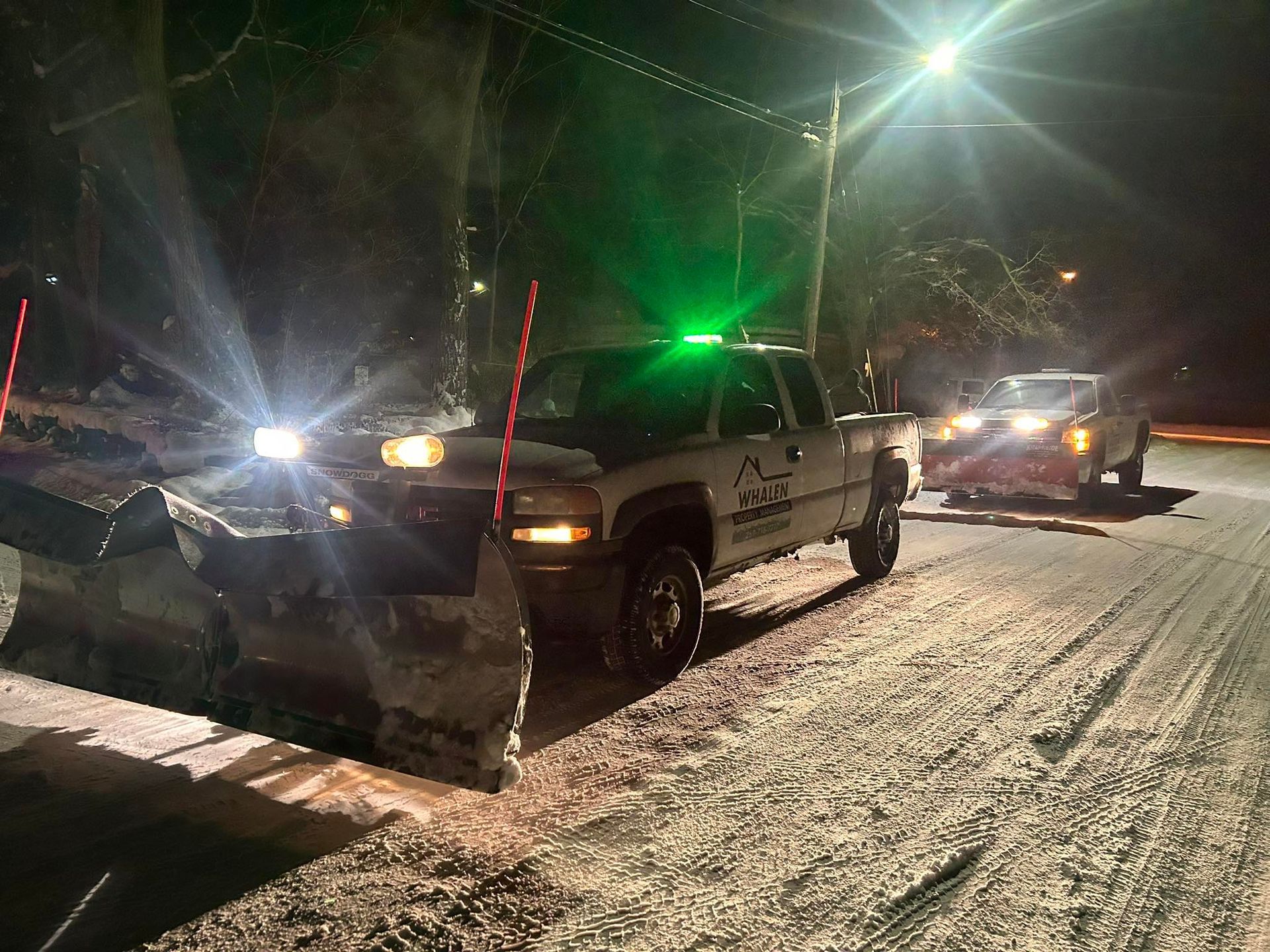 Two snowplow trucks at night with illuminated headlights and green warning lights on a snow-covered, icy road.