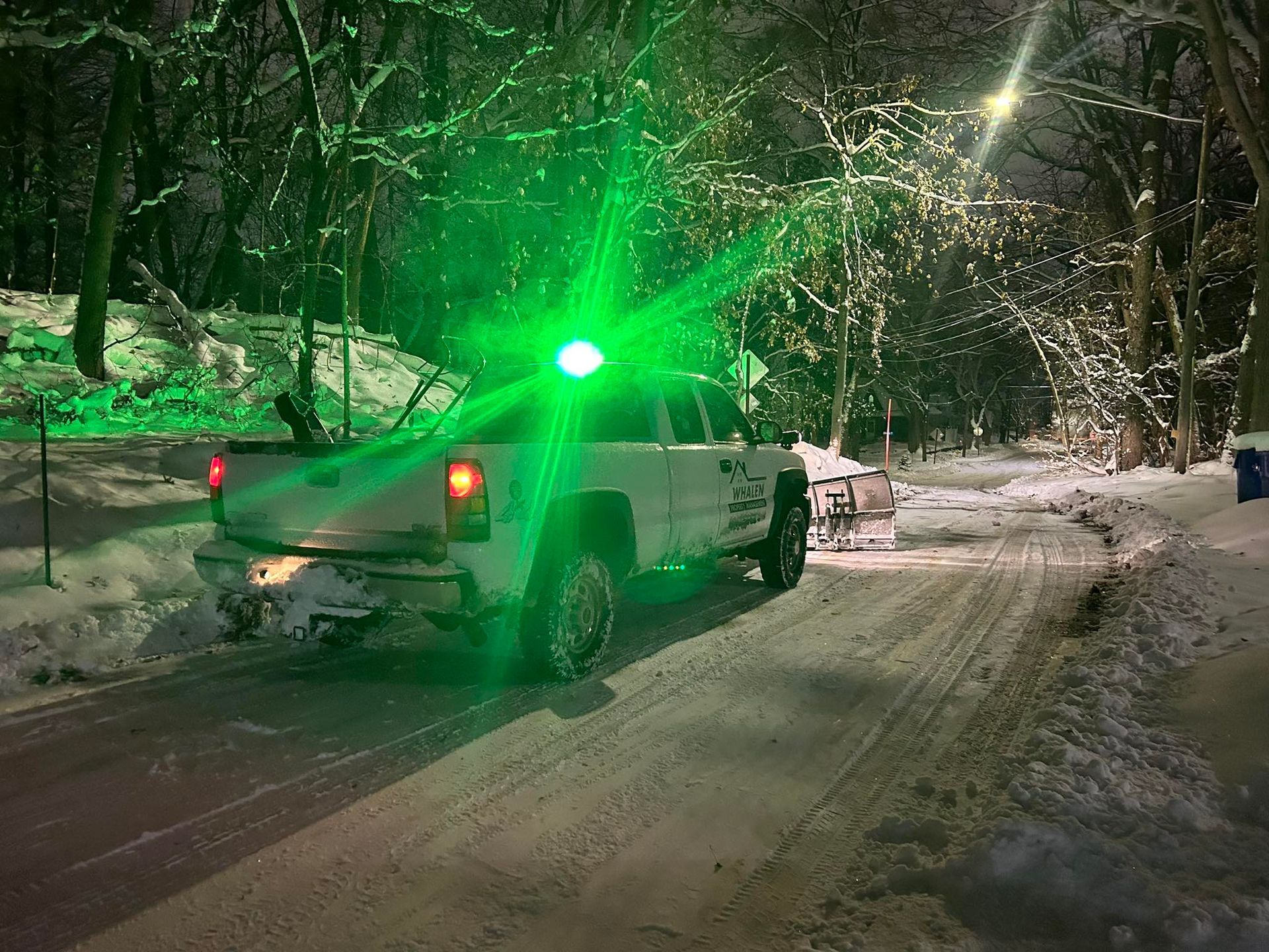 A white pickup truck with a plow on a snow-covered road at night, topped with a bright, glowing green emergency light.