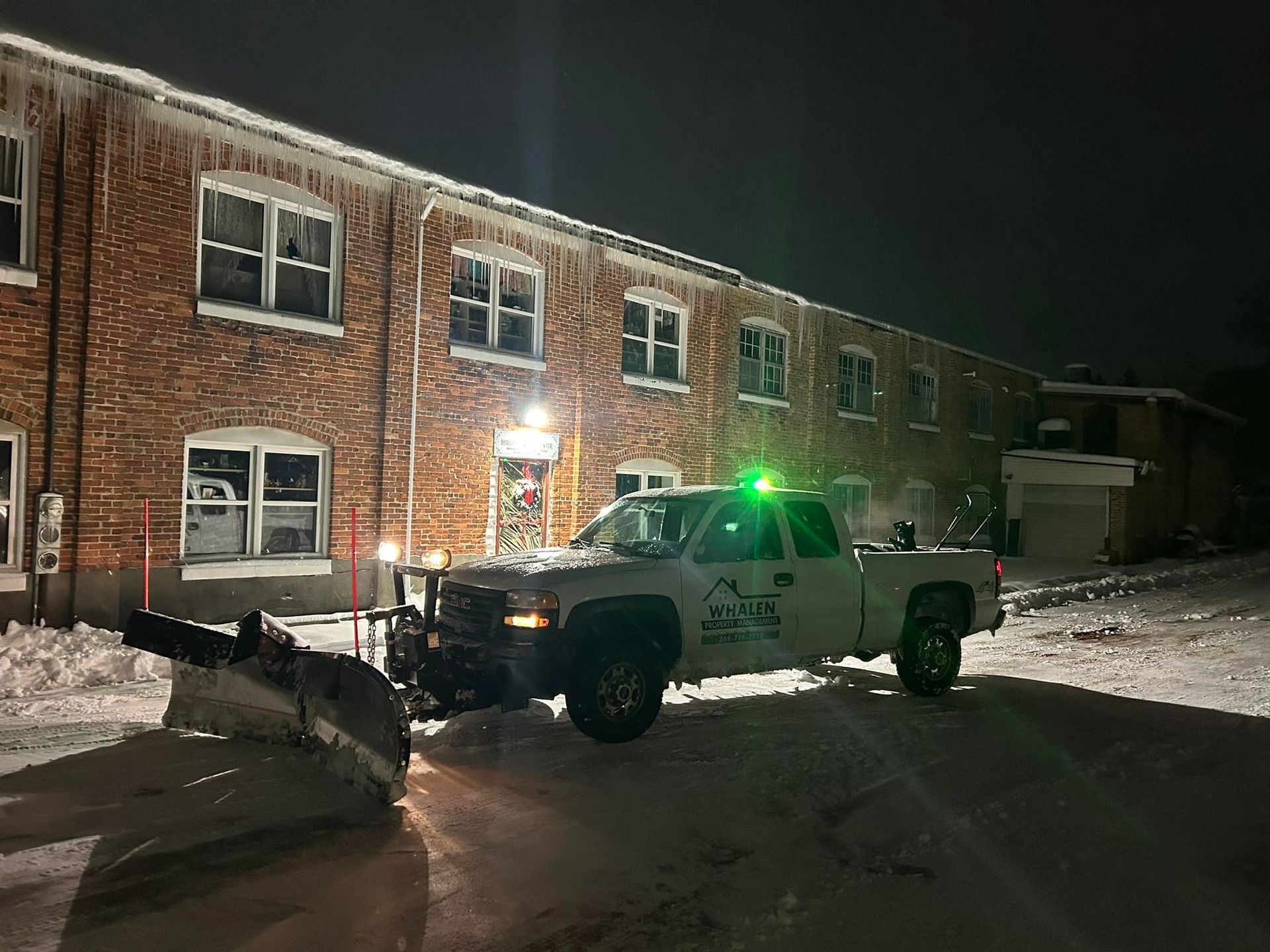 A white snowplow truck with a green light parked at night in front of a multi-story brick building covered in snow.