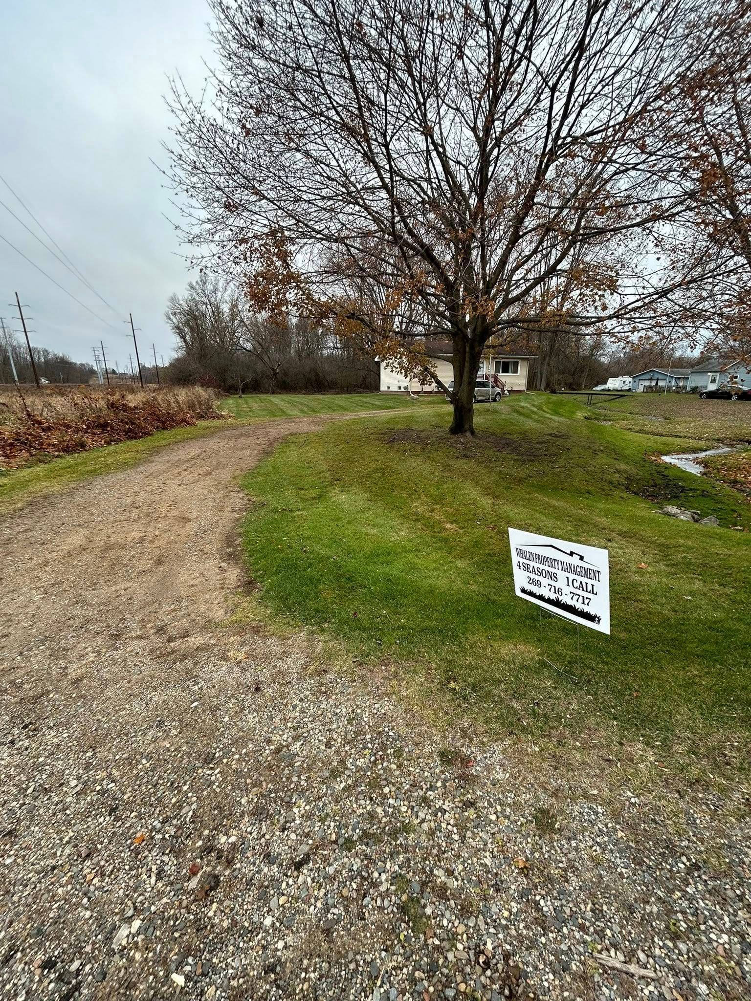 A gravel path leads toward a house behind a bare tree, with a small sign on the grass in the foreground.