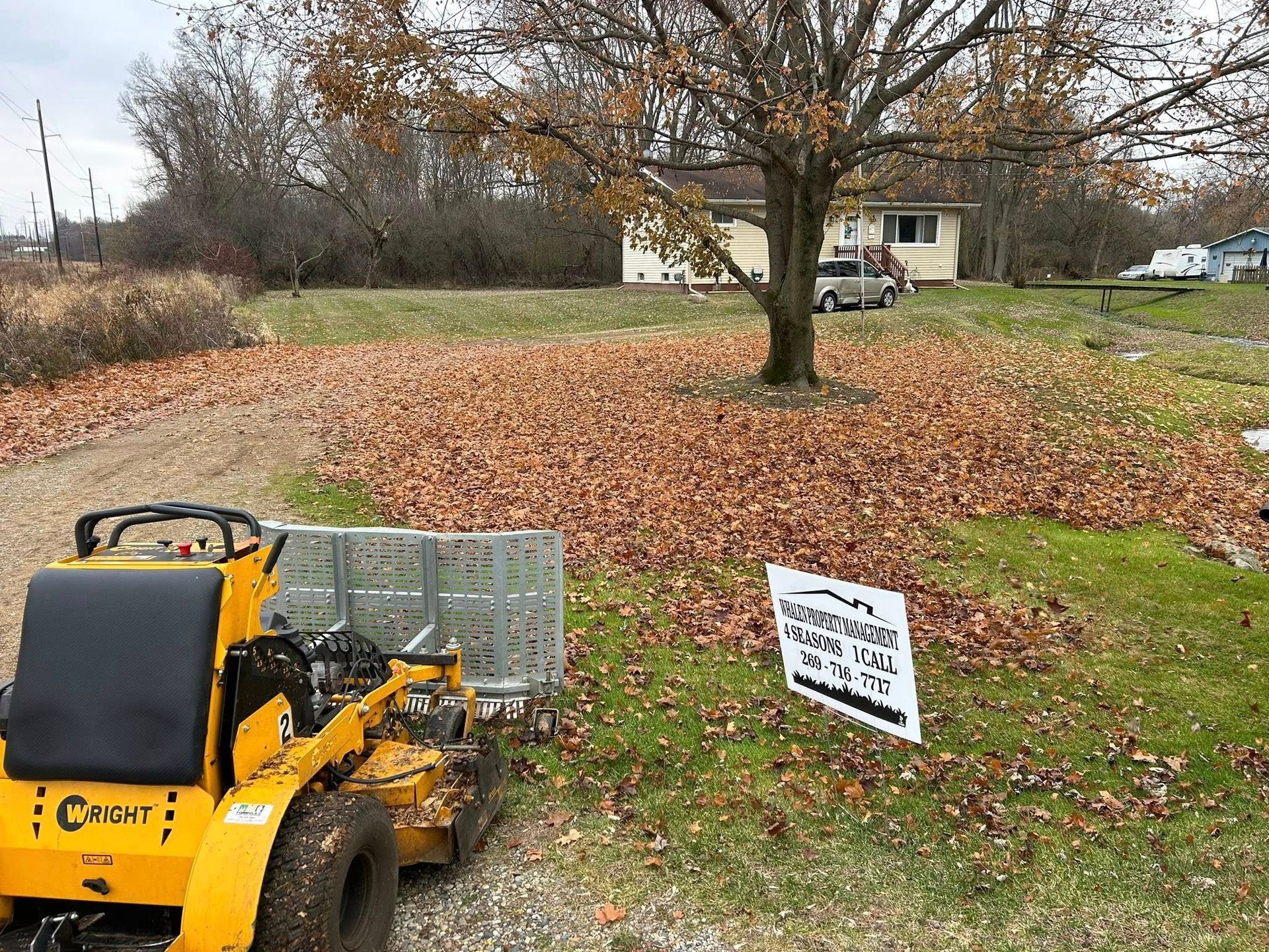 A yellow Wright stand-on lawn mower sits near a sign for a landscaping business on a leaf-covered residential lawn.