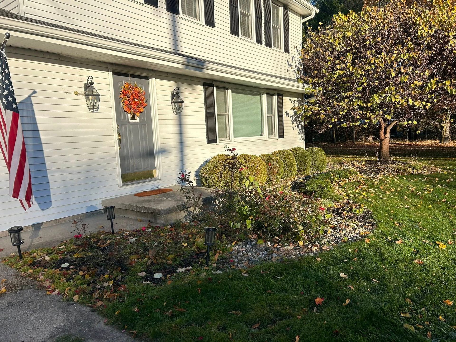 A white two-story house with black shutters, a US flag, an autumn wreath on the door, and a landscaped yard with a tree.