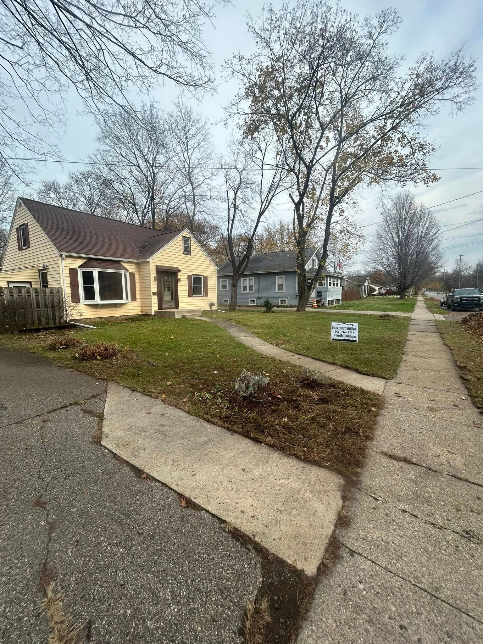 A yellow cottage-style house with a brown roof, large trees, and a sidewalk leading to the front yard.