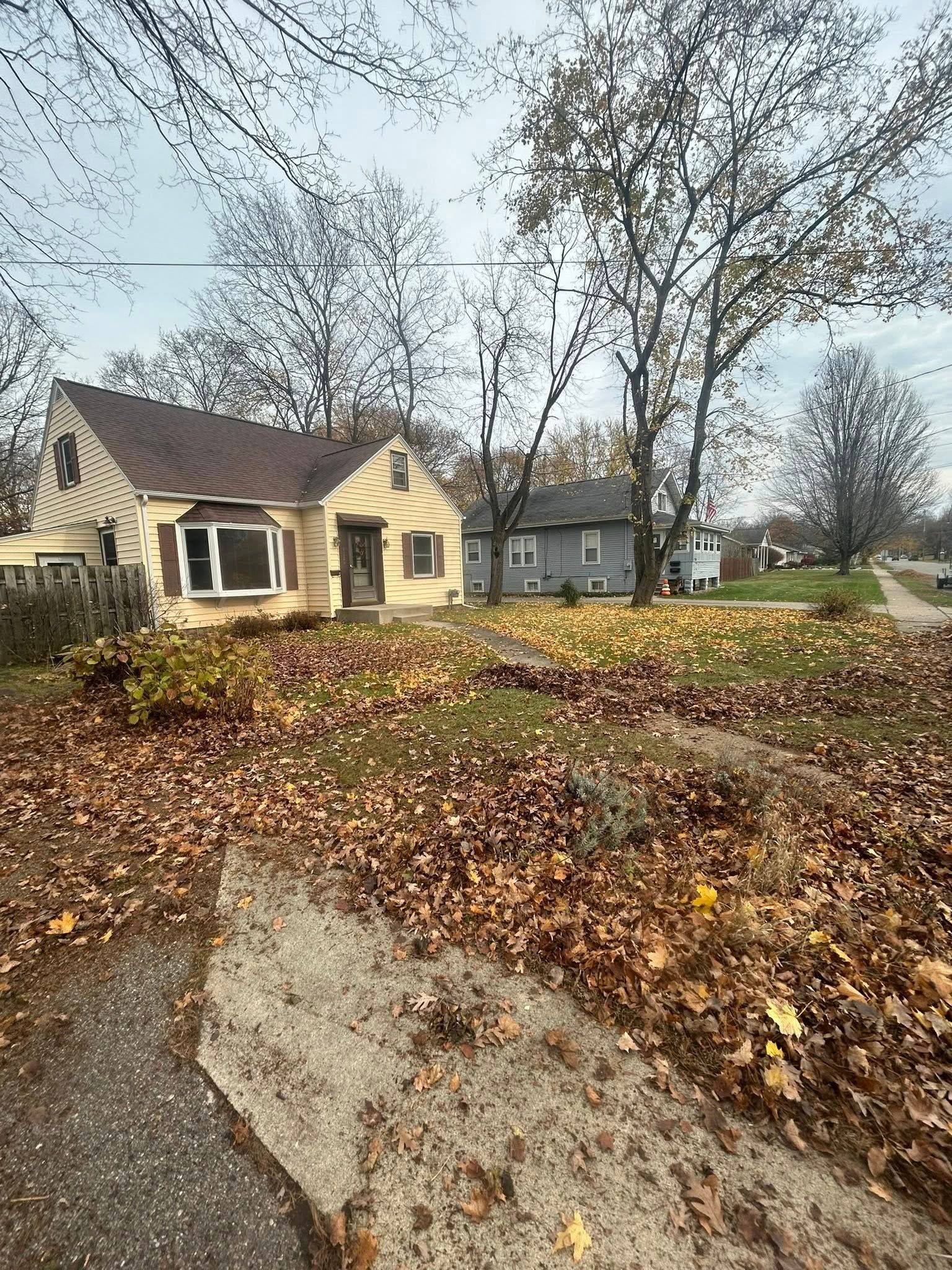 A tan house with a brown roof and a large, leaf-covered yard under a cloudy sky.