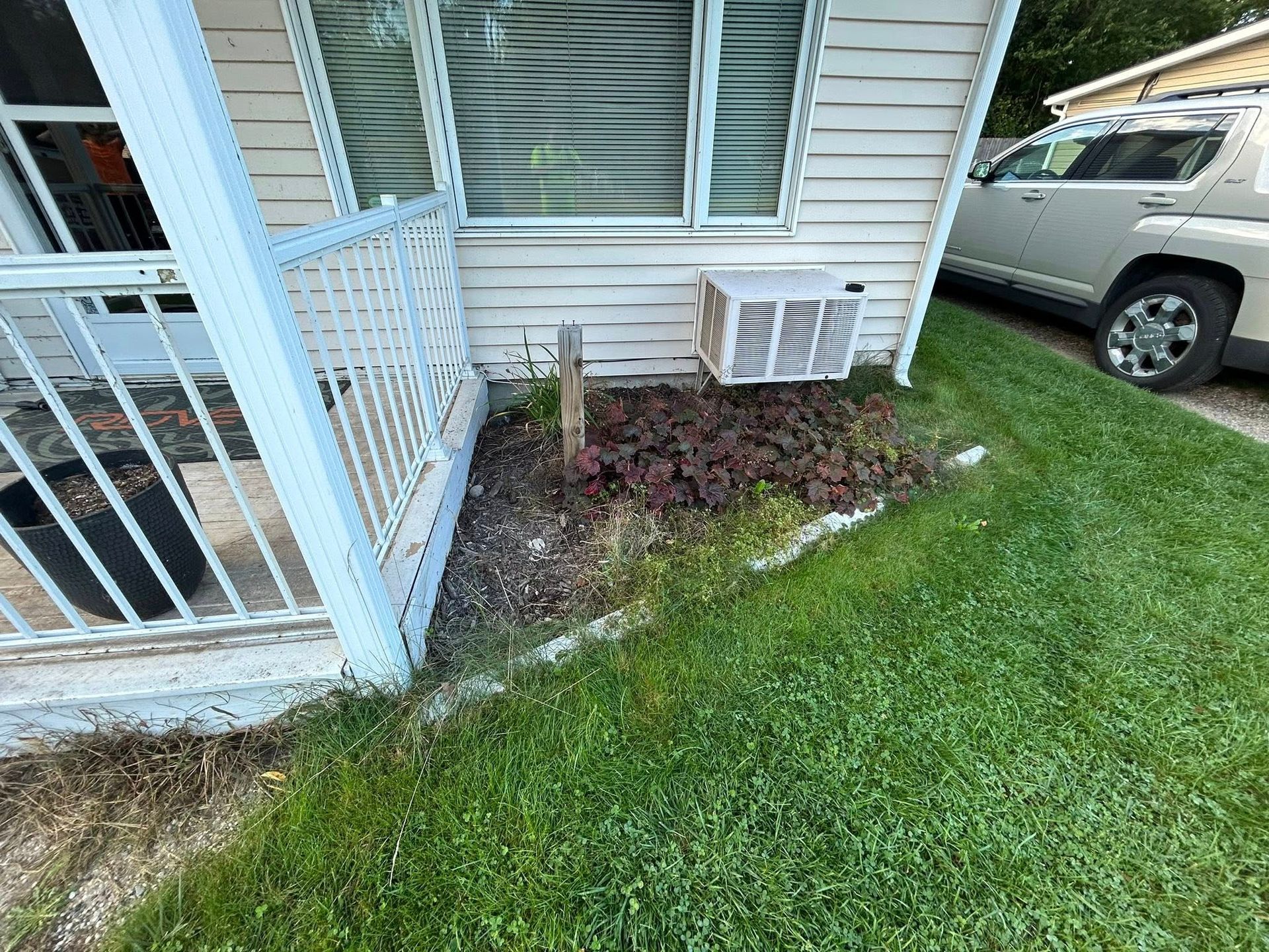 An exterior view of a house corner with a white railing, a window air conditioner, a small garden bed, and a car nearby.