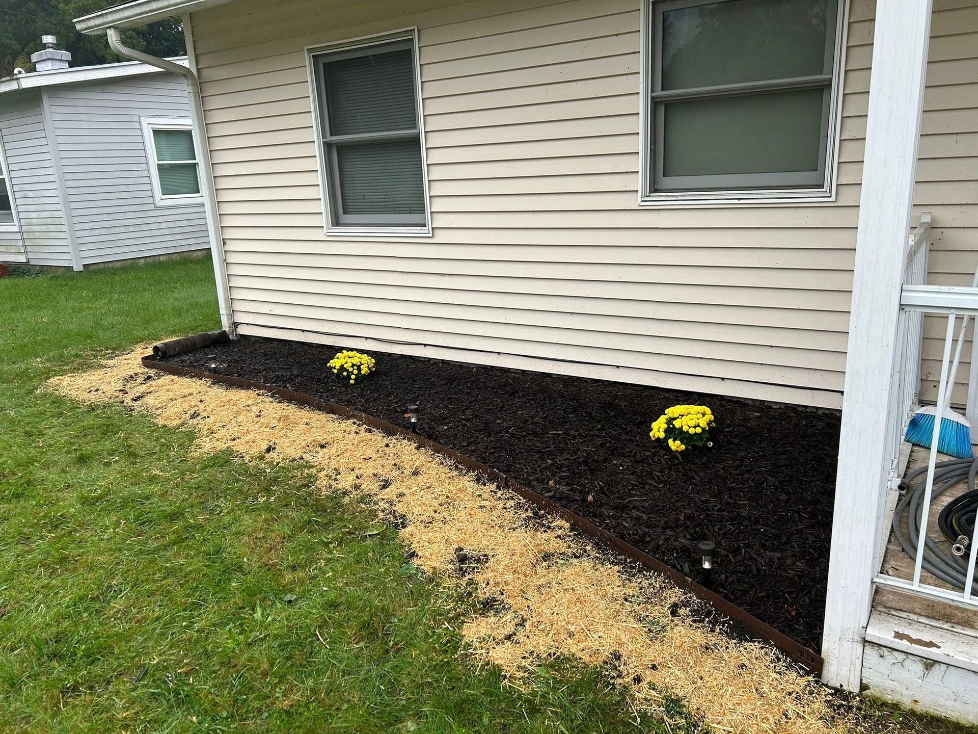A garden bed with dark mulch and two yellow plants against a beige house, with a light tan gravel border along the grass.