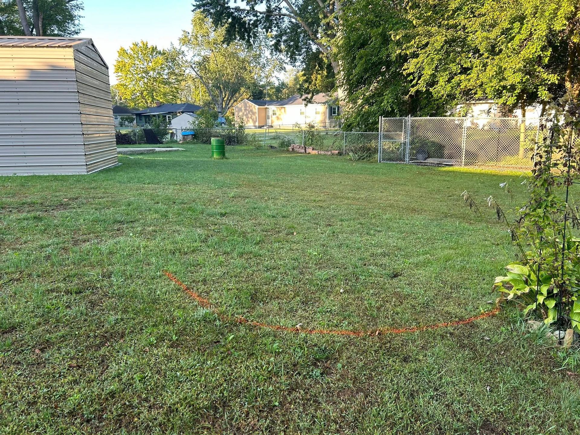 A backyard with a metal shed on the left, an open grassy area, and a fenced garden plot under trees in the distance.
