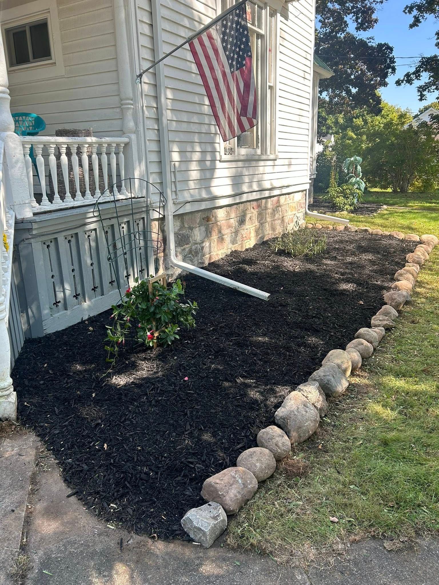 A white house with a porch, a US flag, and a mulched garden bed bordered by stones.