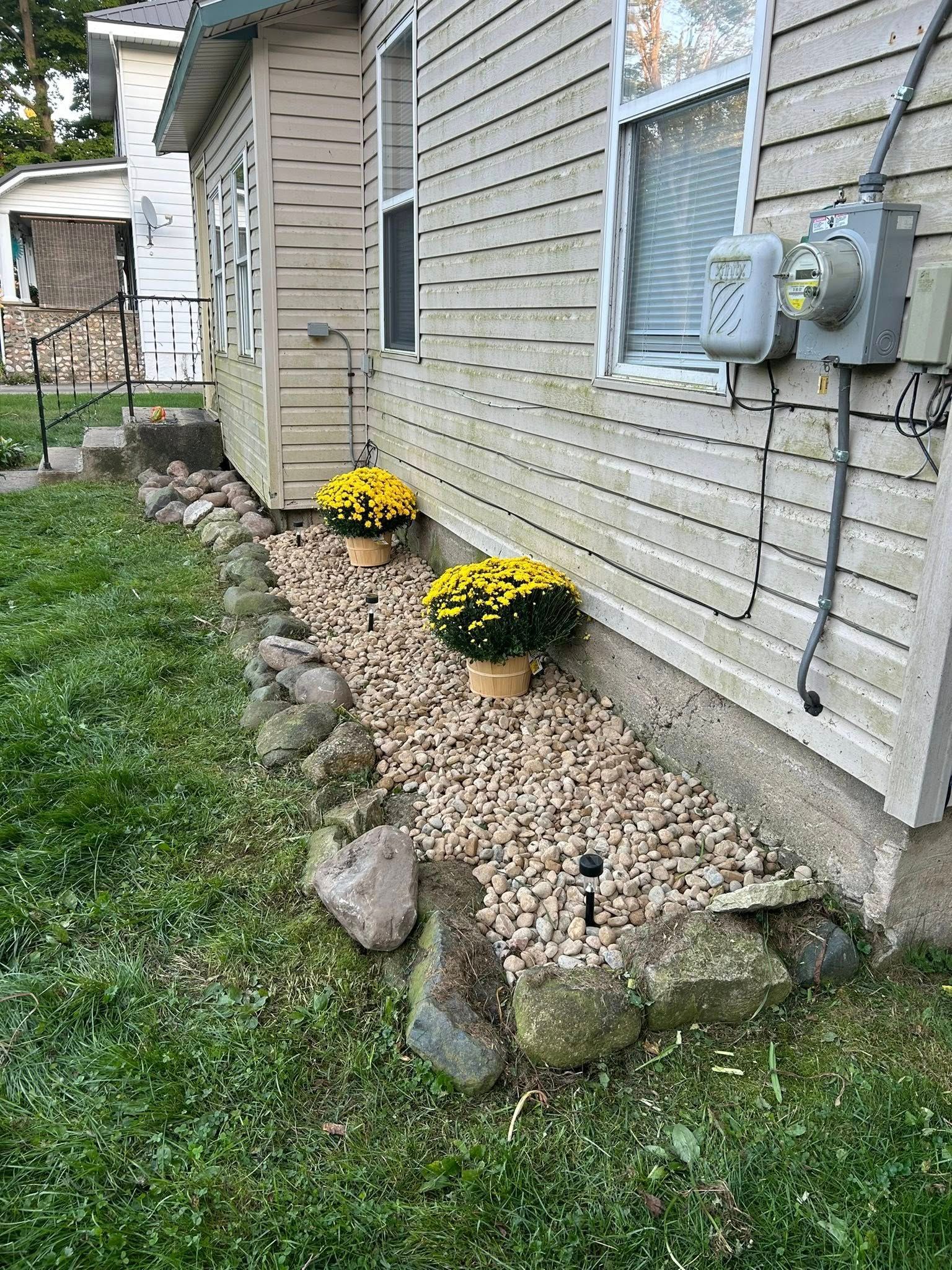 A side view of a house with light beige siding, a stone-lined gravel garden bed, and two pots of yellow chrysanthemums.