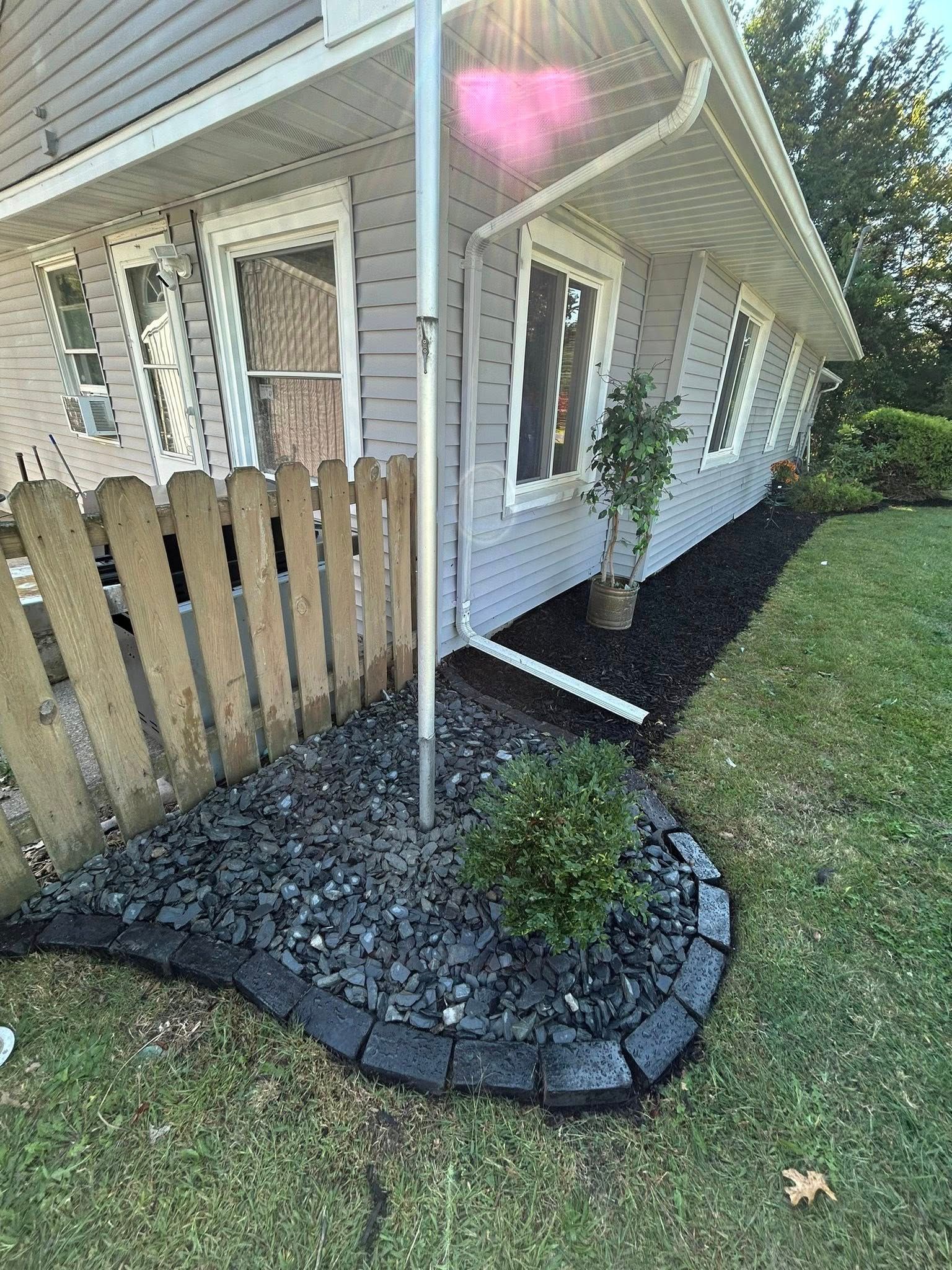 A gray house with a new landscaping bed featuring black mulch, dark gravel, a small shrub, and gray stone edging.