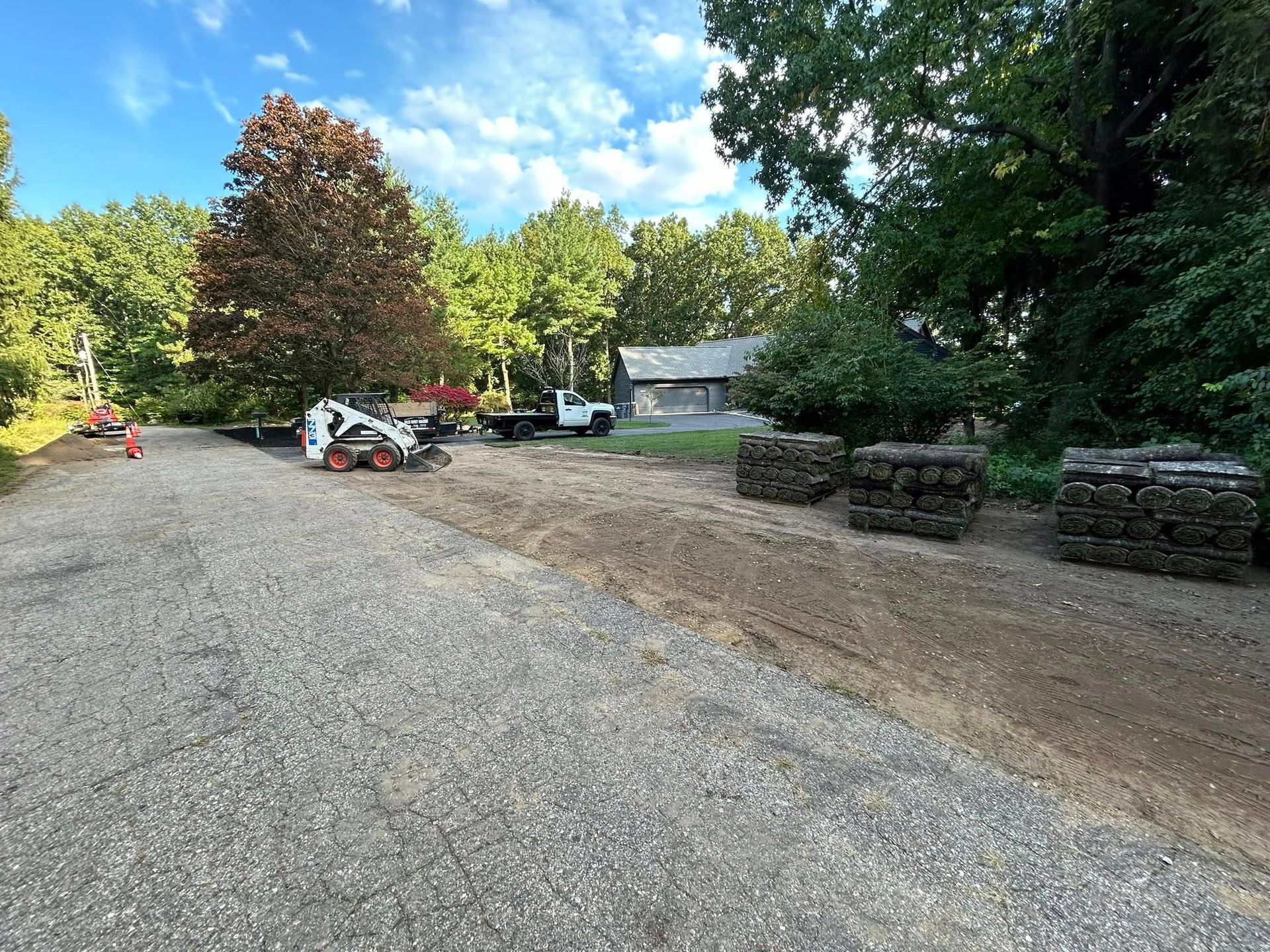 A gravel driveway leads toward a house with a white truck, a skid steer, and stone landscape blocks in a grassy yard.