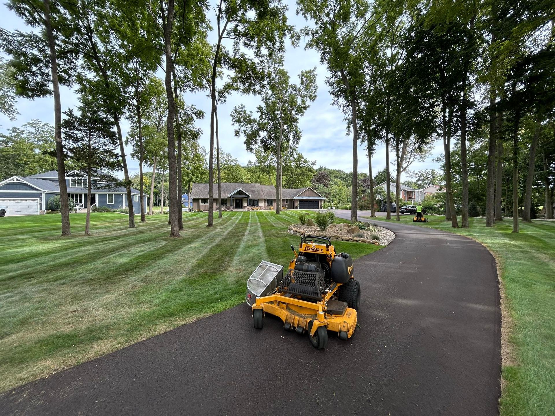 A bright yellow industrial lawn mower sits on a paved driveway in front of a house surrounded by trees and striped grass.