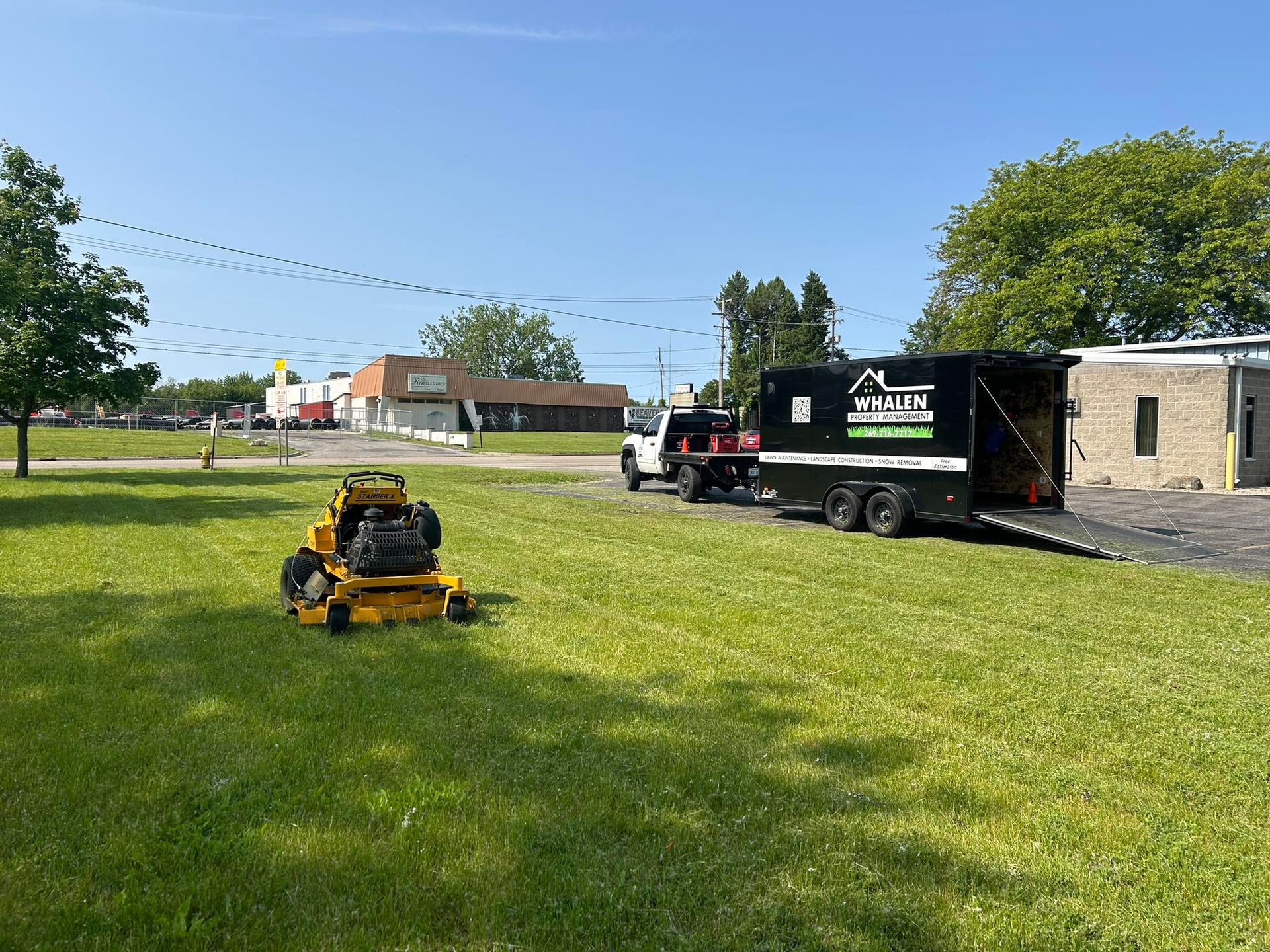 A yellow riding lawn mower sits on a green lawn near a white pickup truck and a black work trailer parked on a gravel lot.