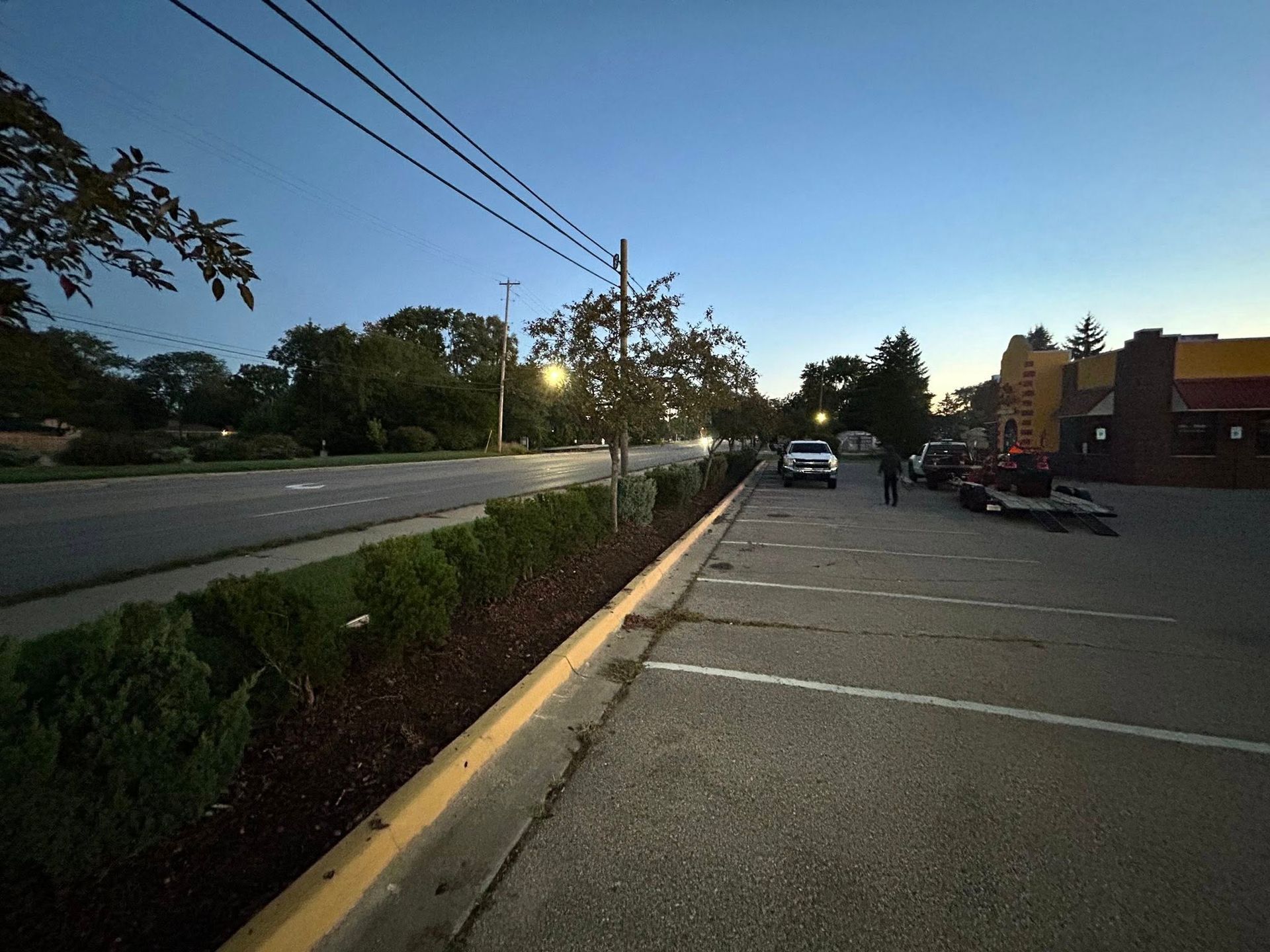 A parking lot at dusk borders a road and green hedge, with a white truck and a person standing near a building.