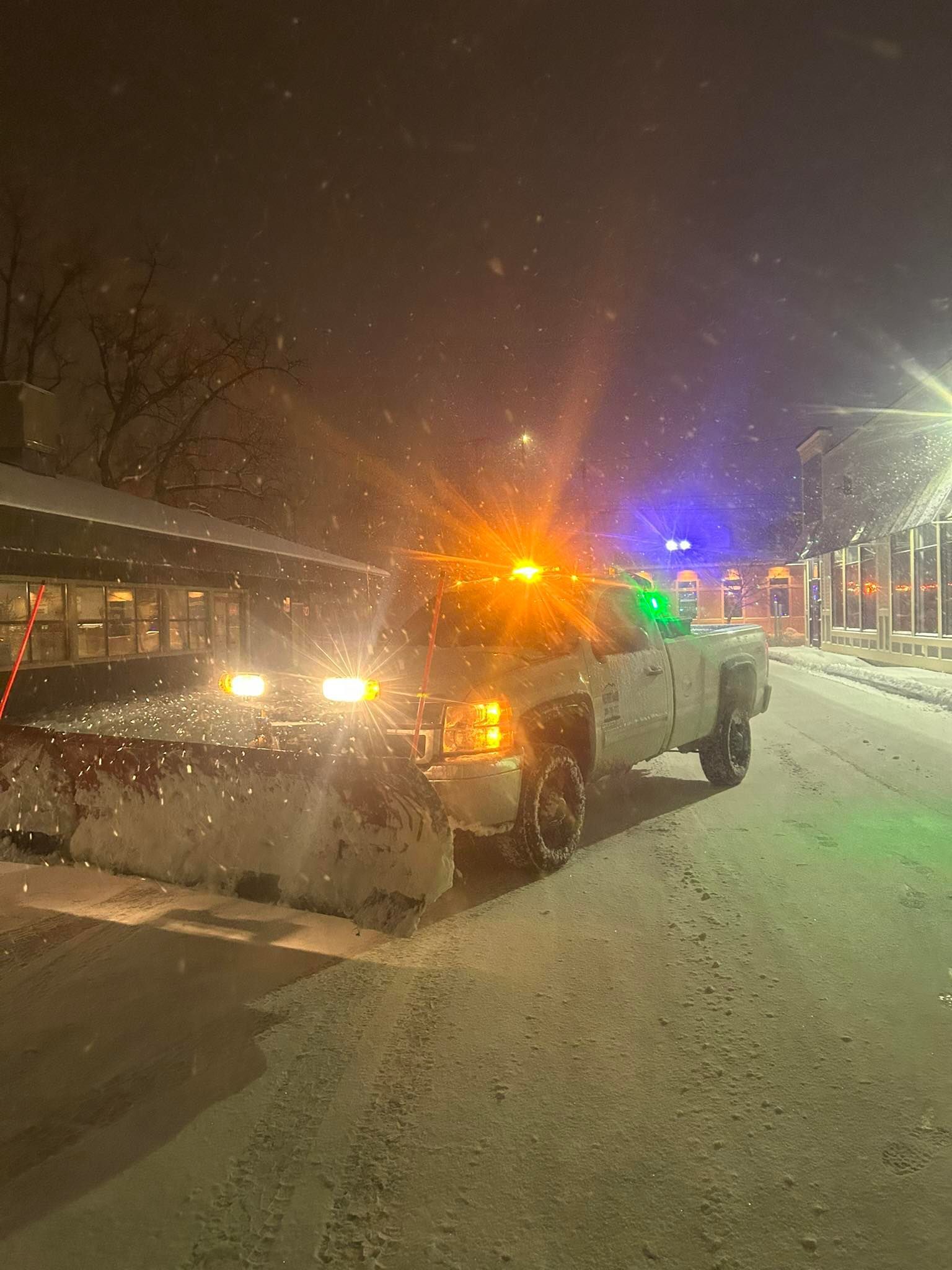A snowplow truck with bright yellow hazard lights clearing snow on a street at night.