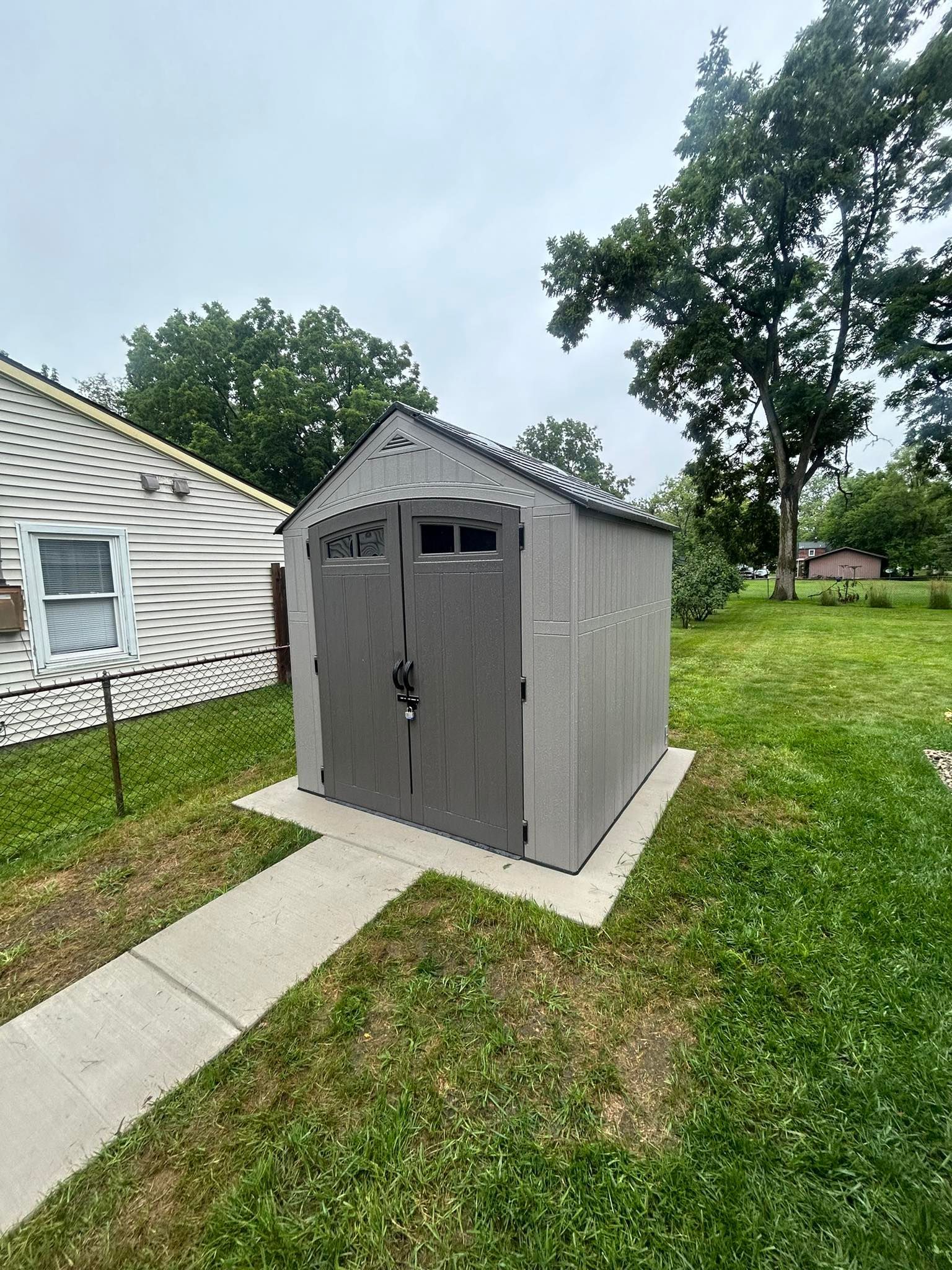 A gray resin storage shed stands on a concrete pad in a grassy yard, next to a white house with horizontal siding.