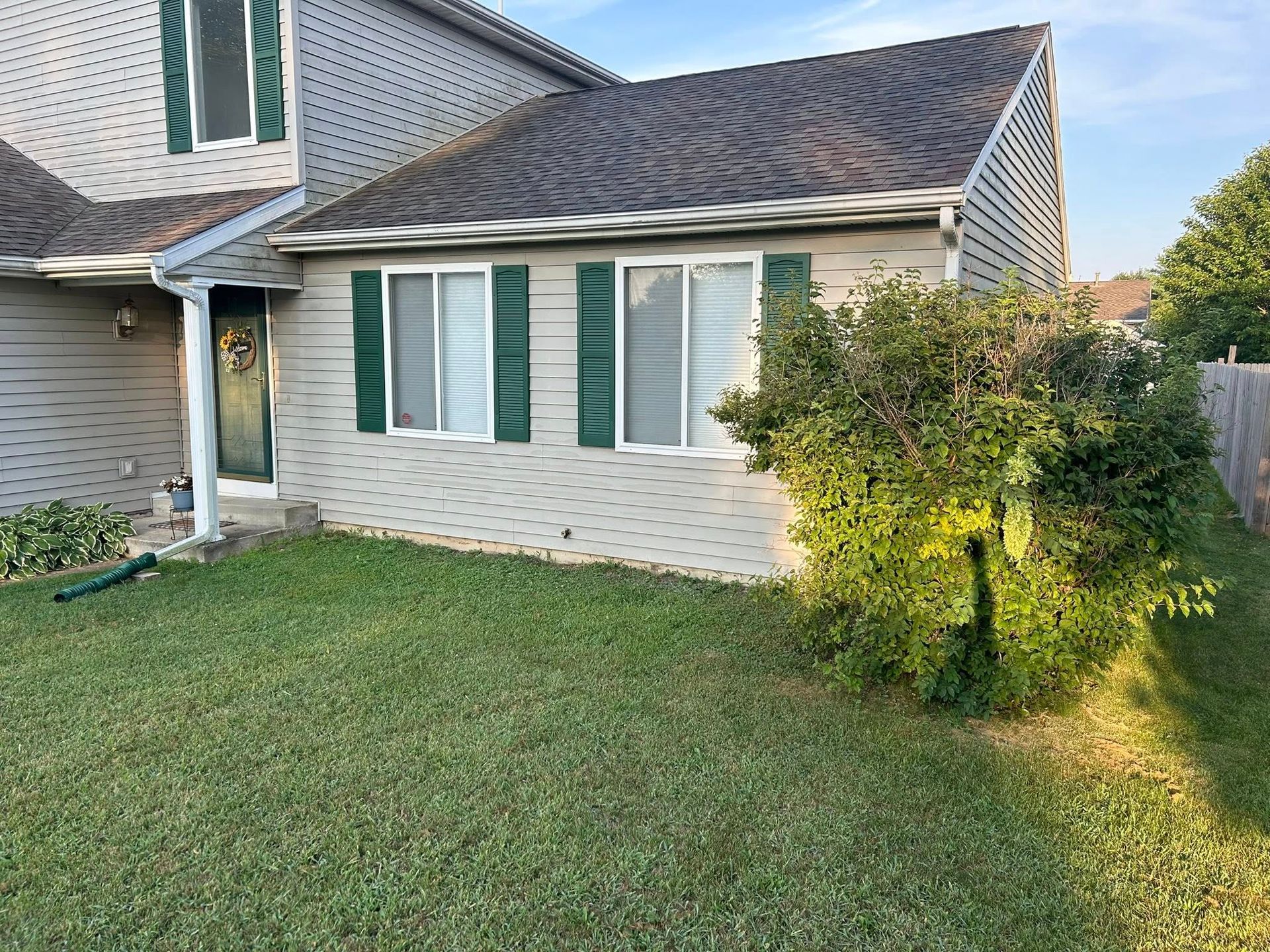 A two-story suburban house with beige siding, dark green shutters, and a large bush in the front yard.
