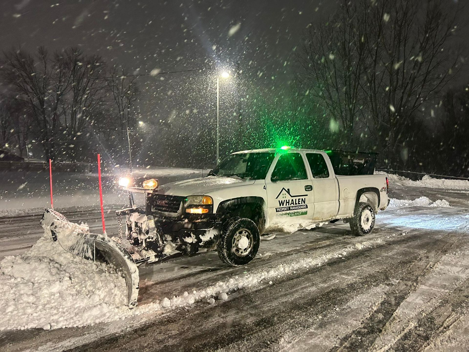 A white pickup truck with a snowplow blade clears snow from a parking lot at night during a winter storm.