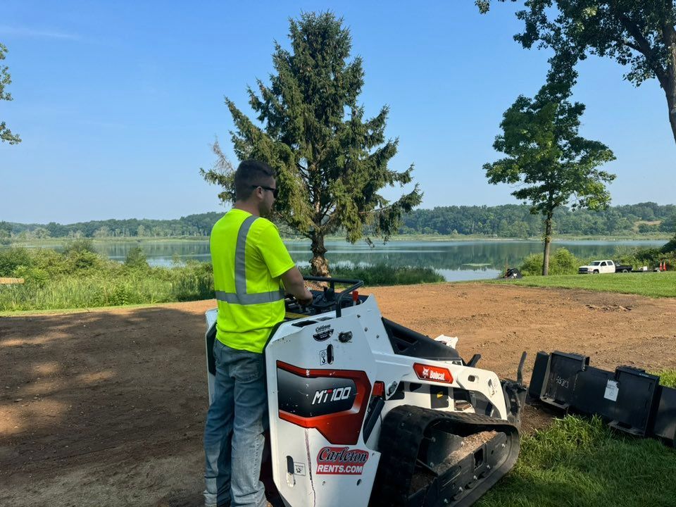 A person in a neon yellow safety vest operating a white Bobcat skid steer on dirt near a lake and trees.