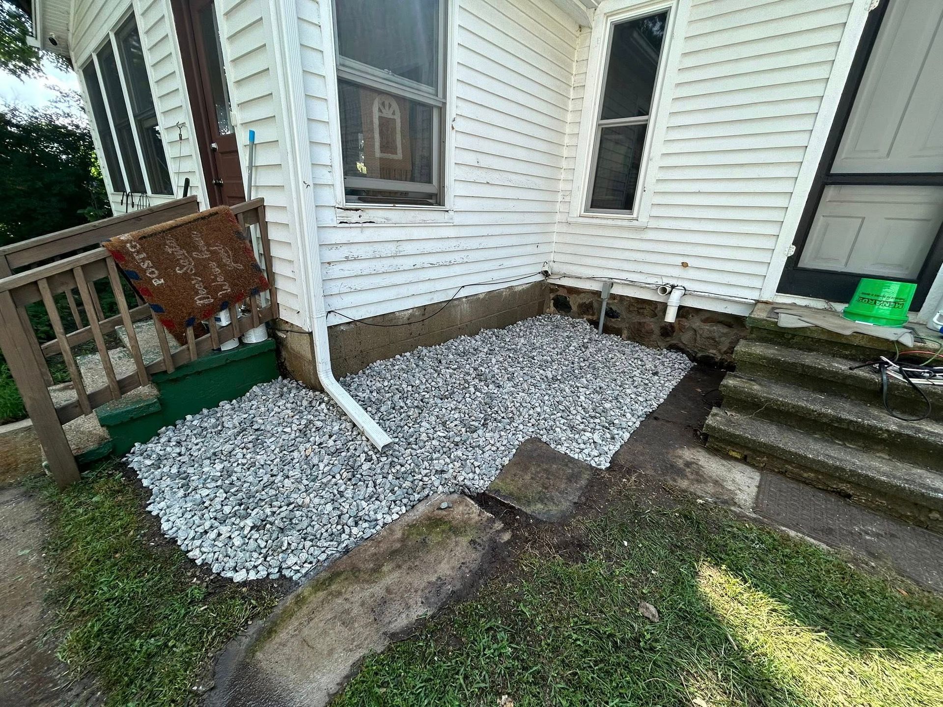 A gravel patch between two sets of concrete house stairs, with a white downspout and white siding on the building.
