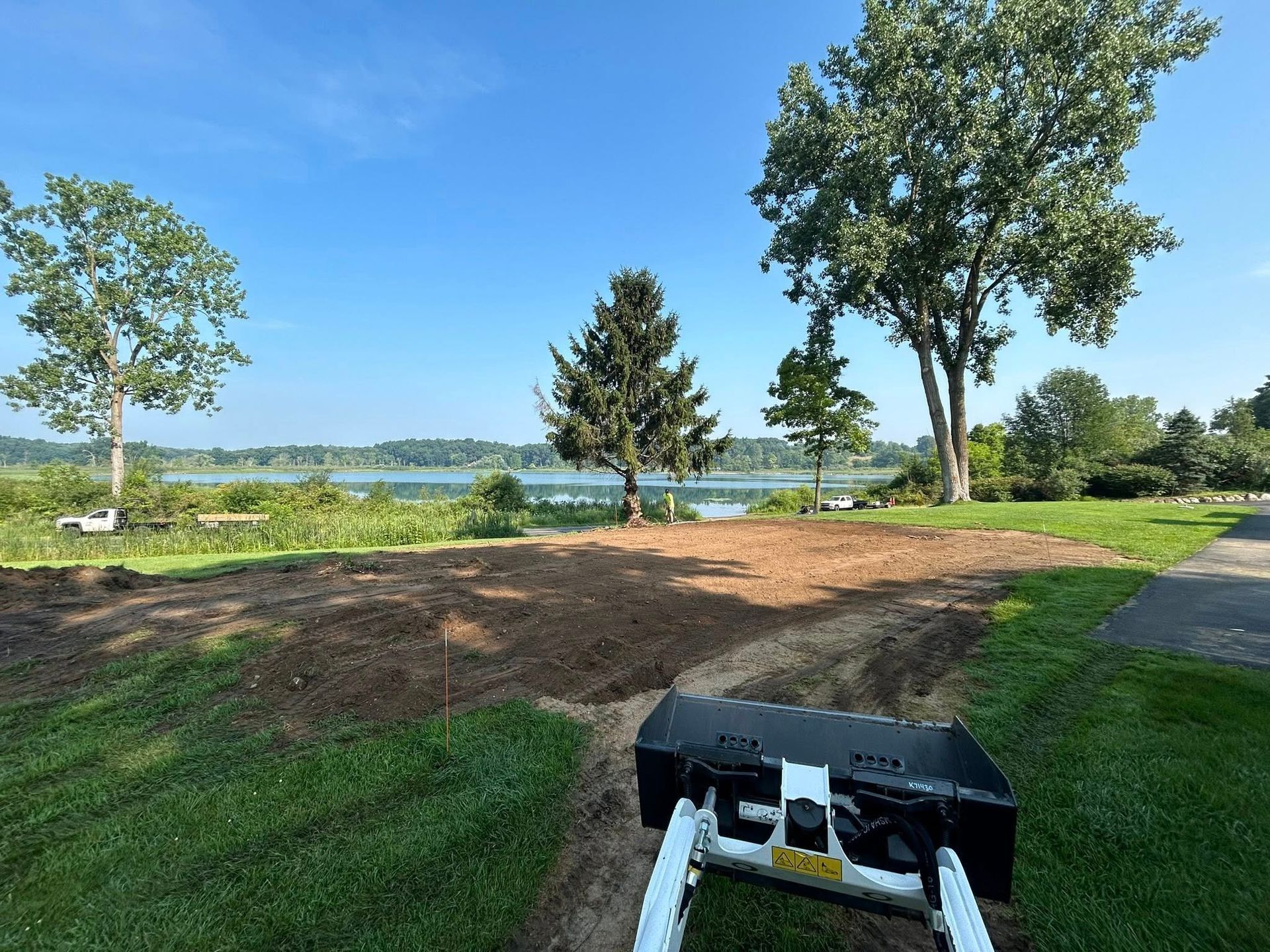 A view from the driver's seat of a skid steer, showing an excavated dirt lot overlooking a lake on a sunny day.