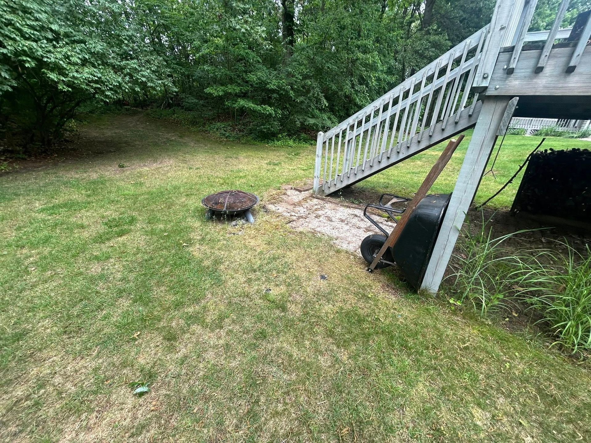 A backyard lawn featuring a fire pit, a gravel path under a wooden deck staircase, and a black wheelbarrow.