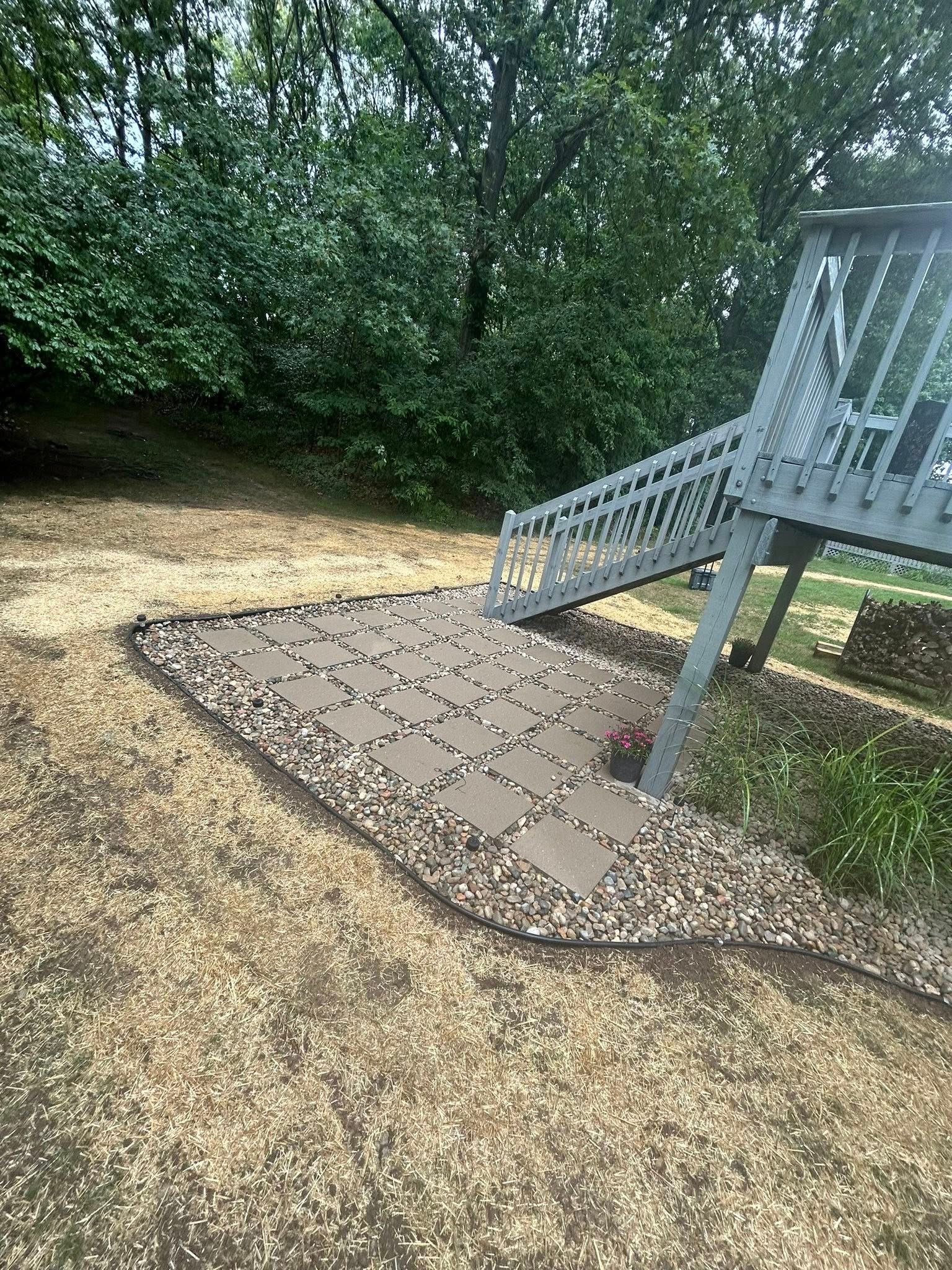 A light gray wooden deck staircase leading down to a small, square stone paver patio surrounded by gravel in a yard.