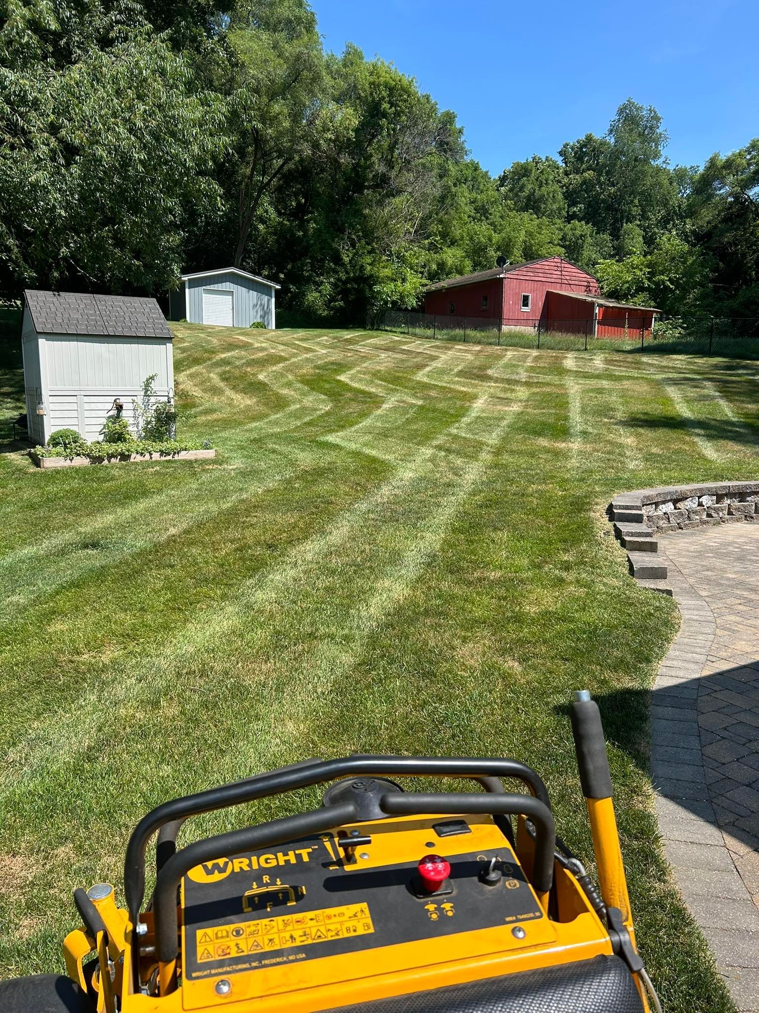 A bright yellow Wright lawn mower overlooks a freshly mowed backyard with a gray shed, a small garage, and a red barn.