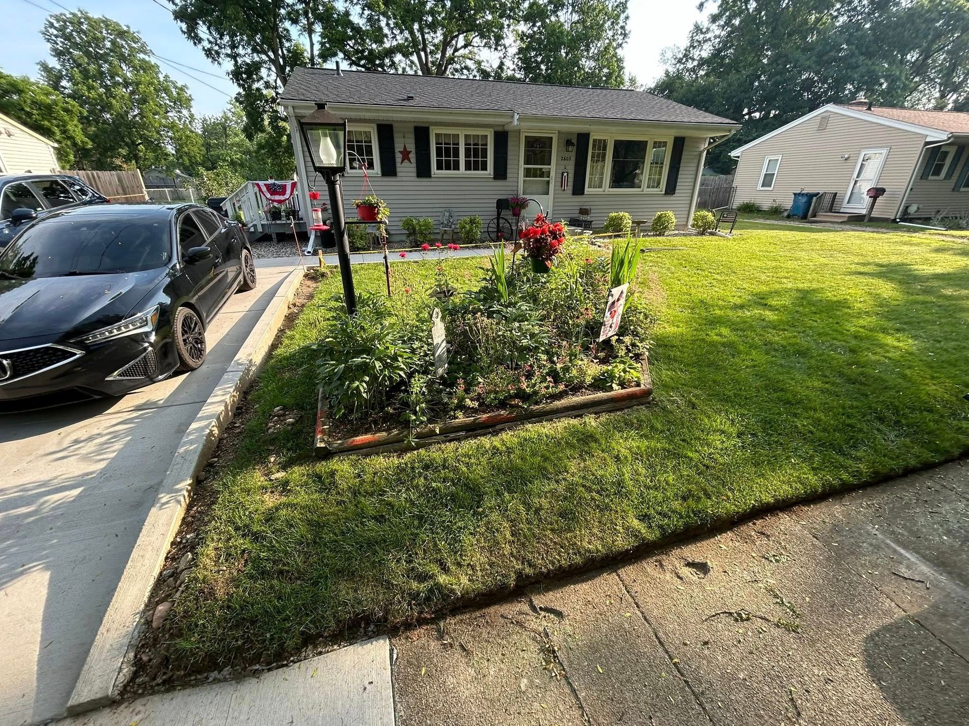 A gray house with a lawn, a central flower bed, and a black car parked in the driveway.
