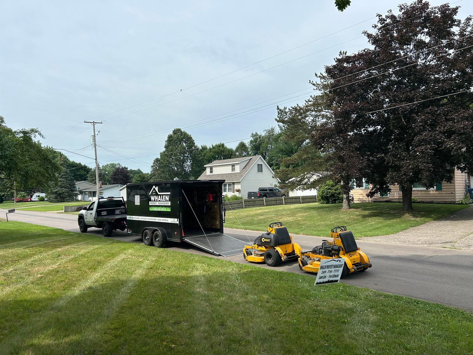 A white truck towing a black landscaping trailer with two yellow ride-on lawnmowers parked on a residential street.