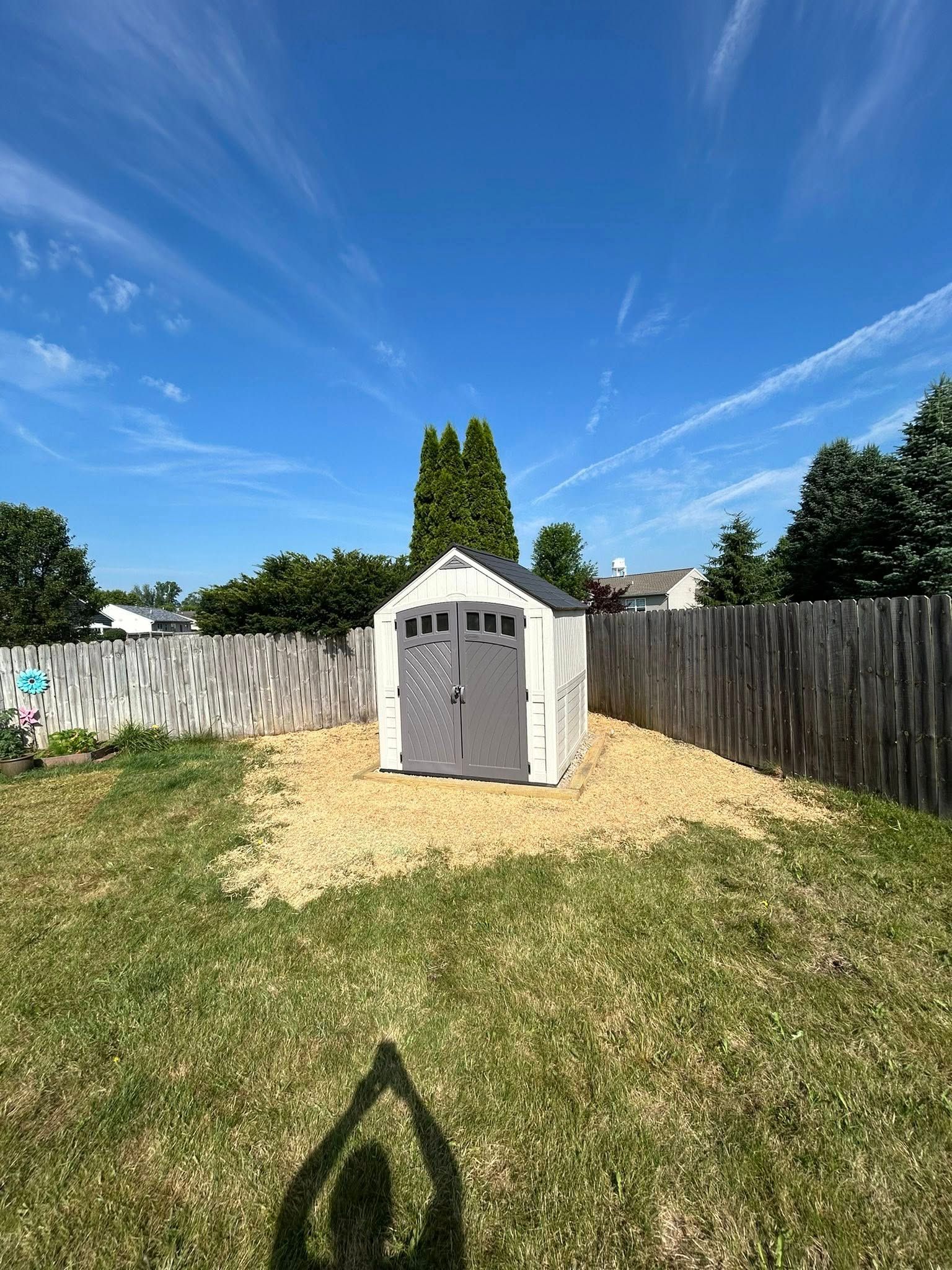 A white garden shed with grey doors sits on a patch of wood chips in a backyard under a clear blue sky.