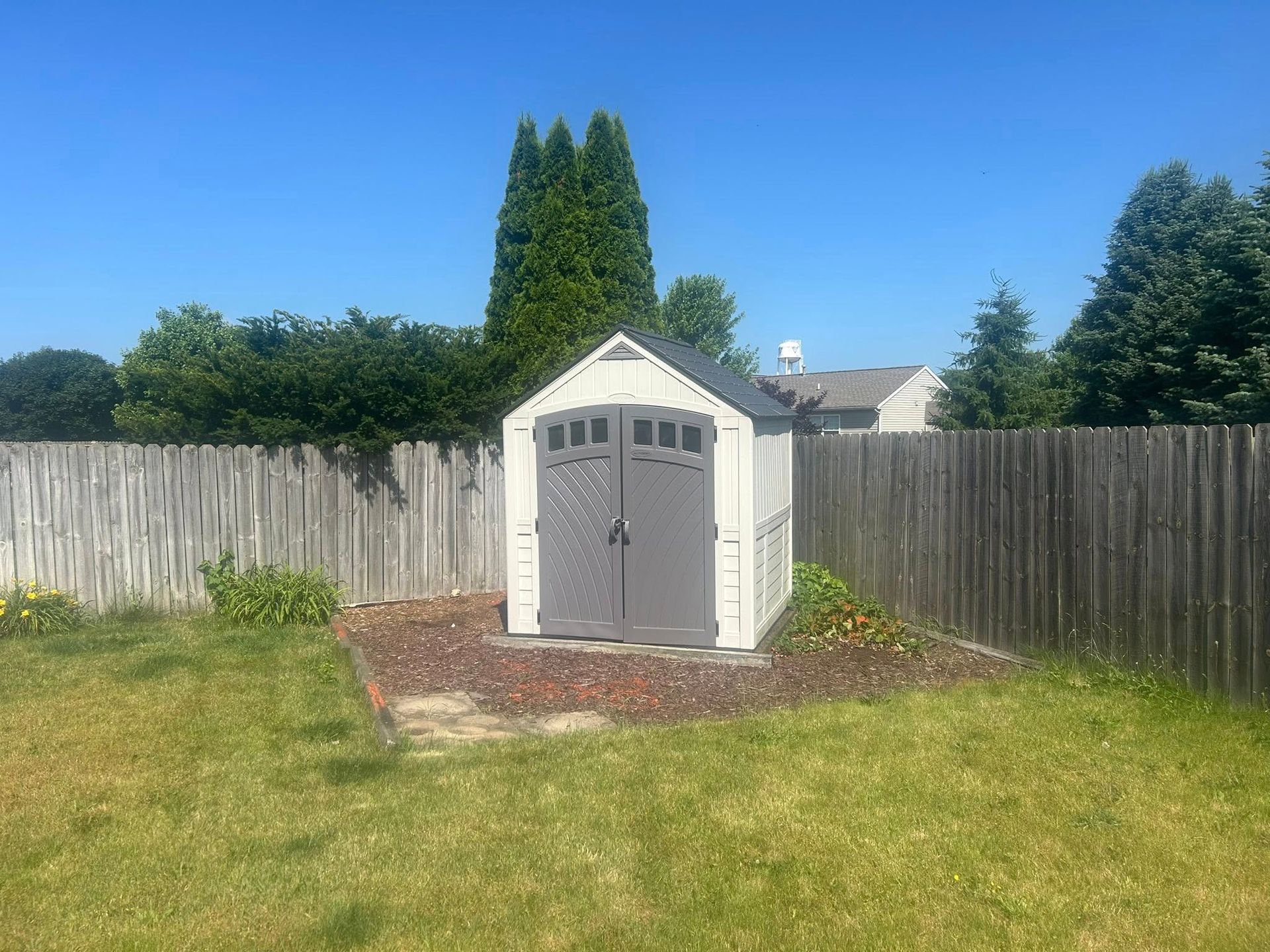 A grey and white shed sits on a gravel patch in a backyard, surrounded by a wooden fence and green lawn under a blue sky.