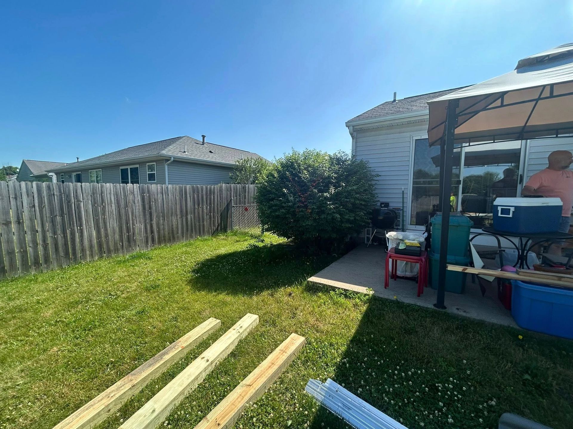 A backyard patio with a gazebo, outdoor furniture, a cooler, and wooden lumber resting on the lawn near a wooden fence.