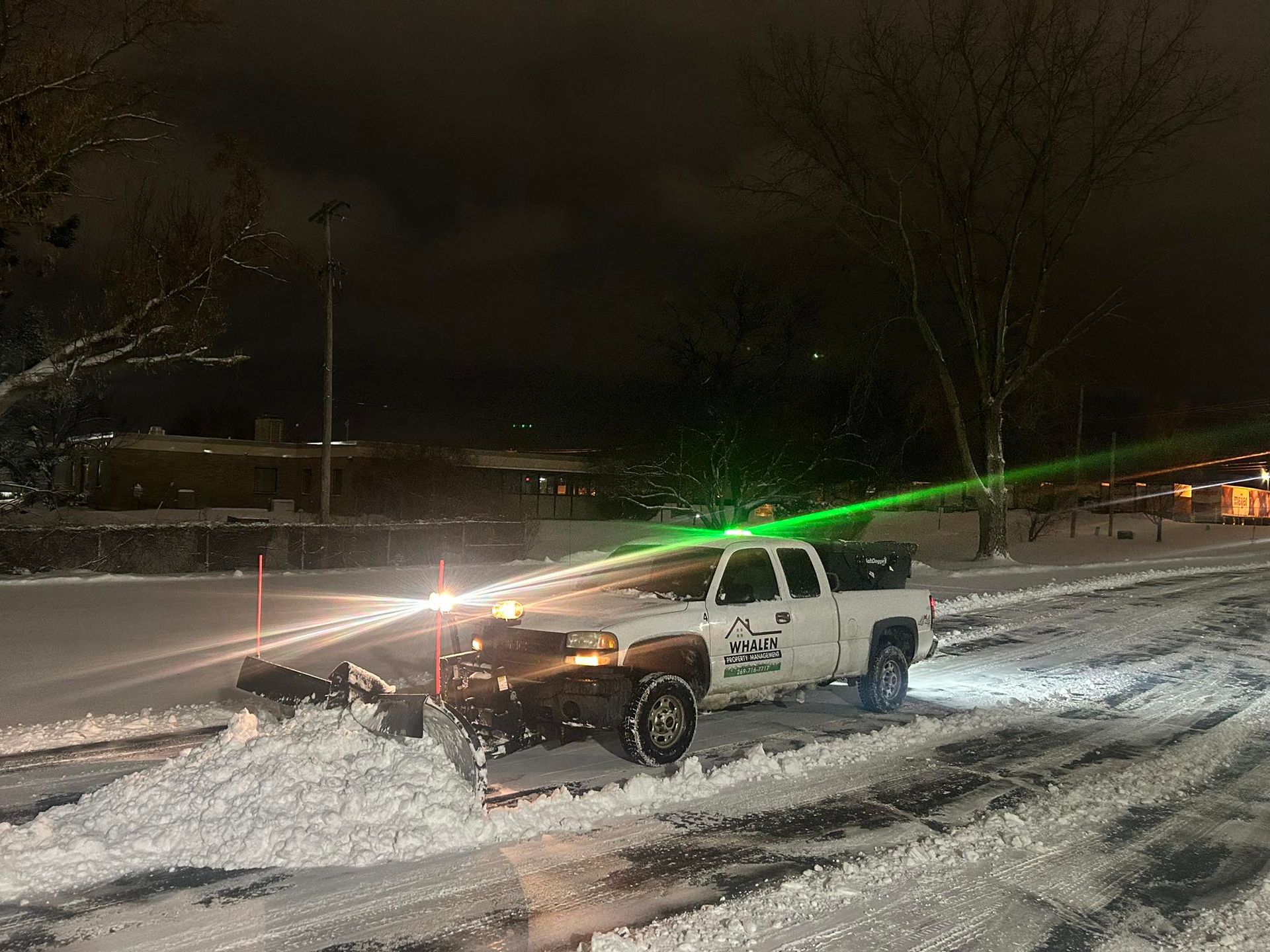 A white snowplow truck clearing snow from a parking lot at night.