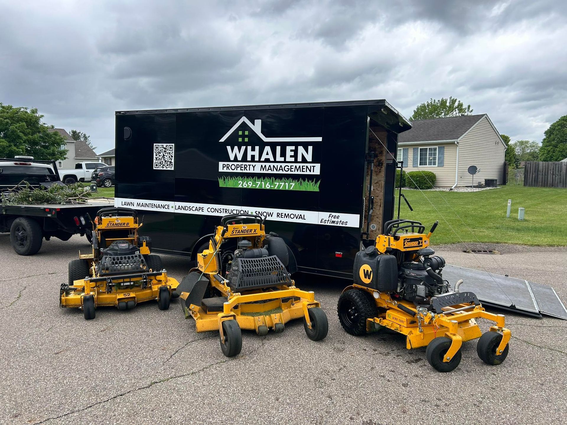 Three yellow commercial lawn mowers parked in front of a black trailer for Whalen Property Maintenance on an asphalt lot.