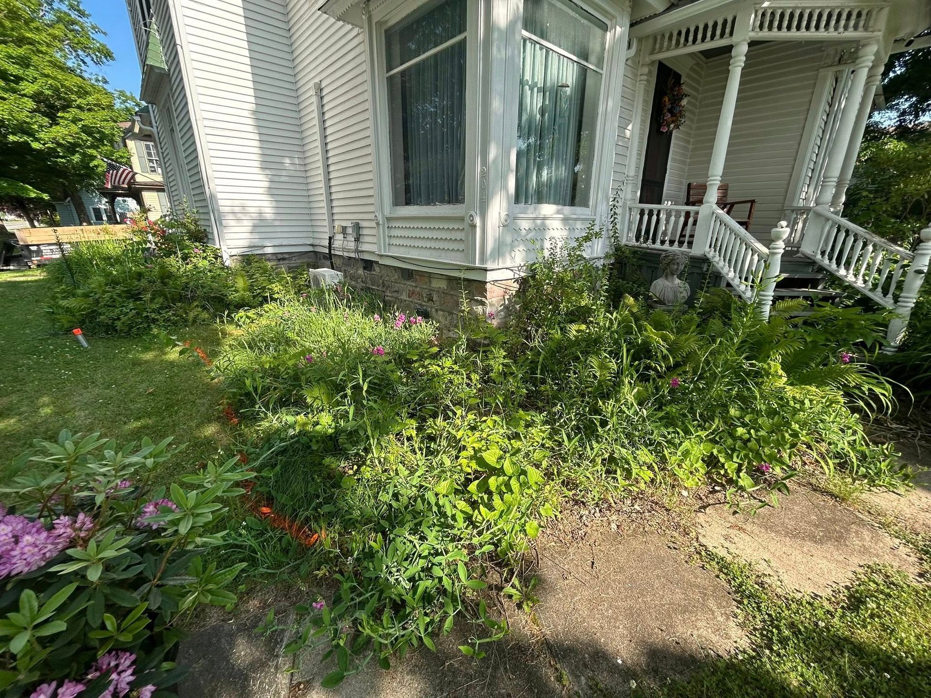 A white Victorian-style house features a bay window and a covered front porch with ornate railings, set above a garden.