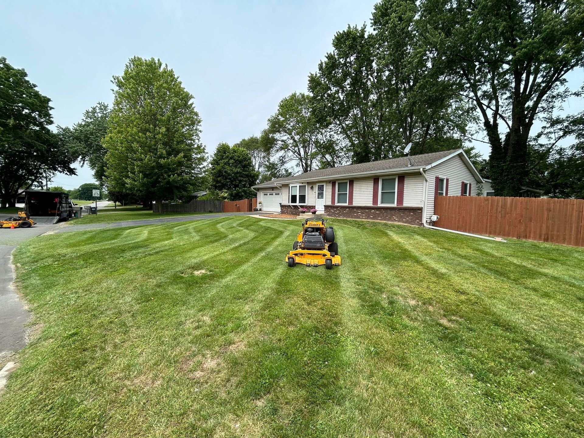 A yellow commercial mower sits on a freshly mowed front lawn with striped grass in front of a white house with a fence.