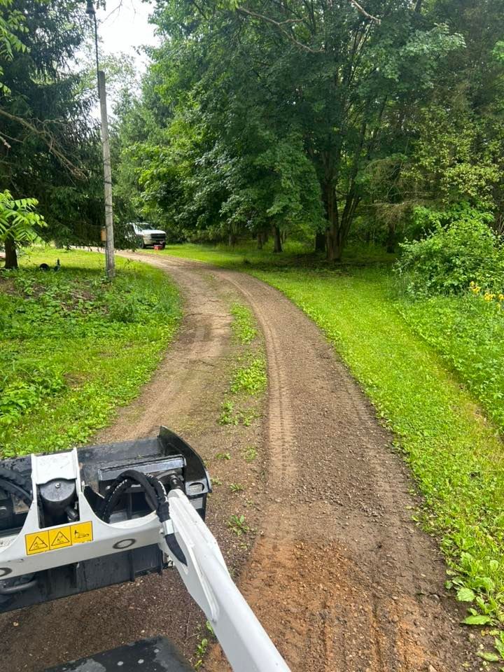 View from the operator's seat of a white skid-steer loader parked on a gravel path leading through a wooded area.