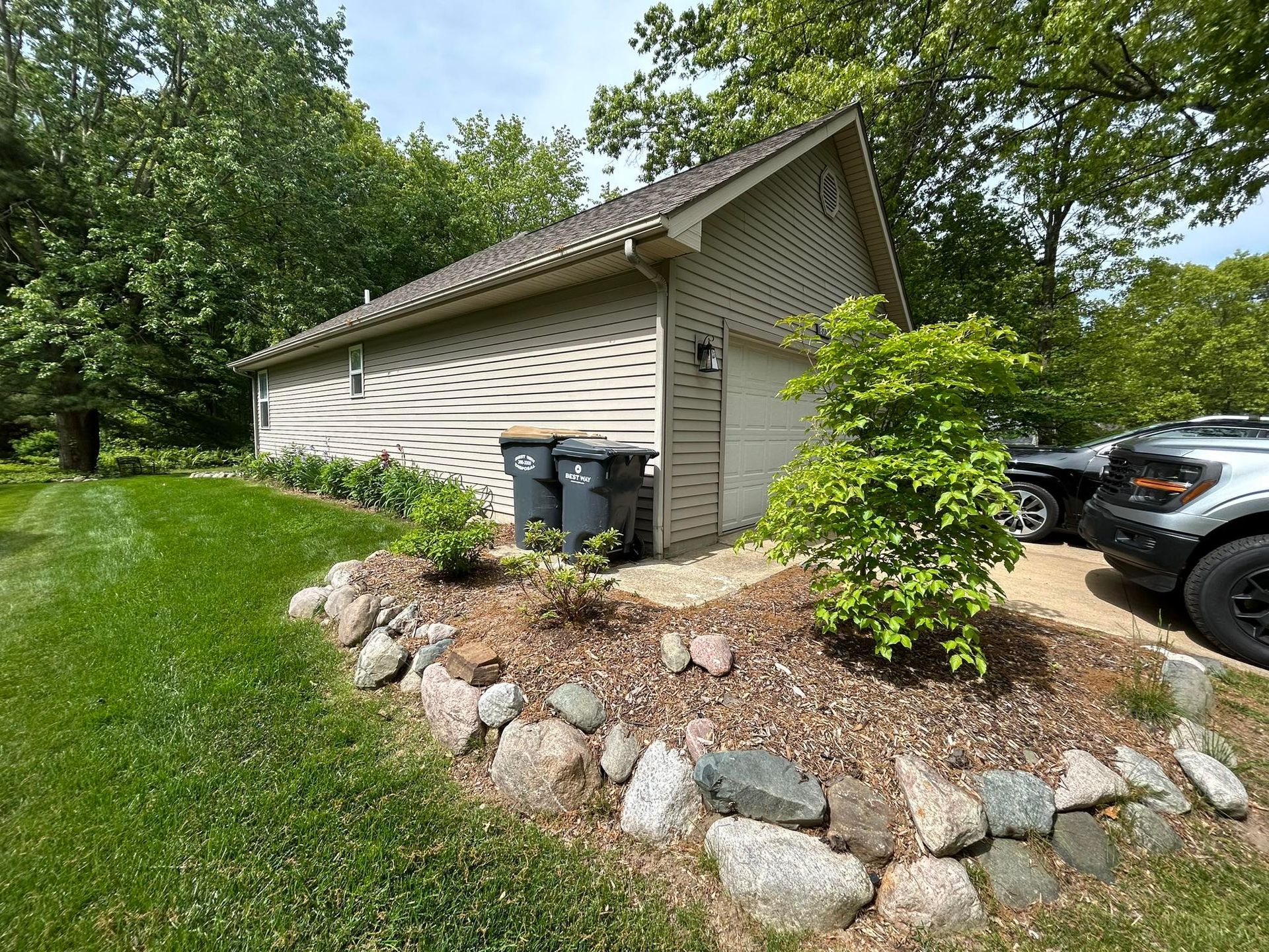 A tan detached garage with a rock-bordered garden bed in front, set against green trees and a car on the right.