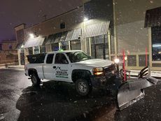 A white pickup truck with a snow plow attached parked in front of a building during a night snowstorm.