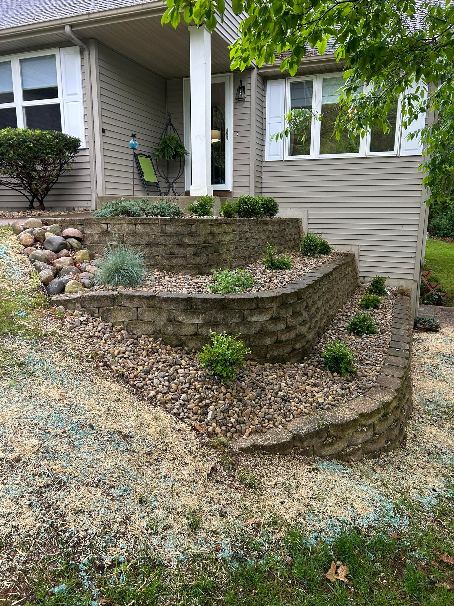 A tan, single-story house with tiered stone retaining walls featuring small green shrubs and landscaping stones.