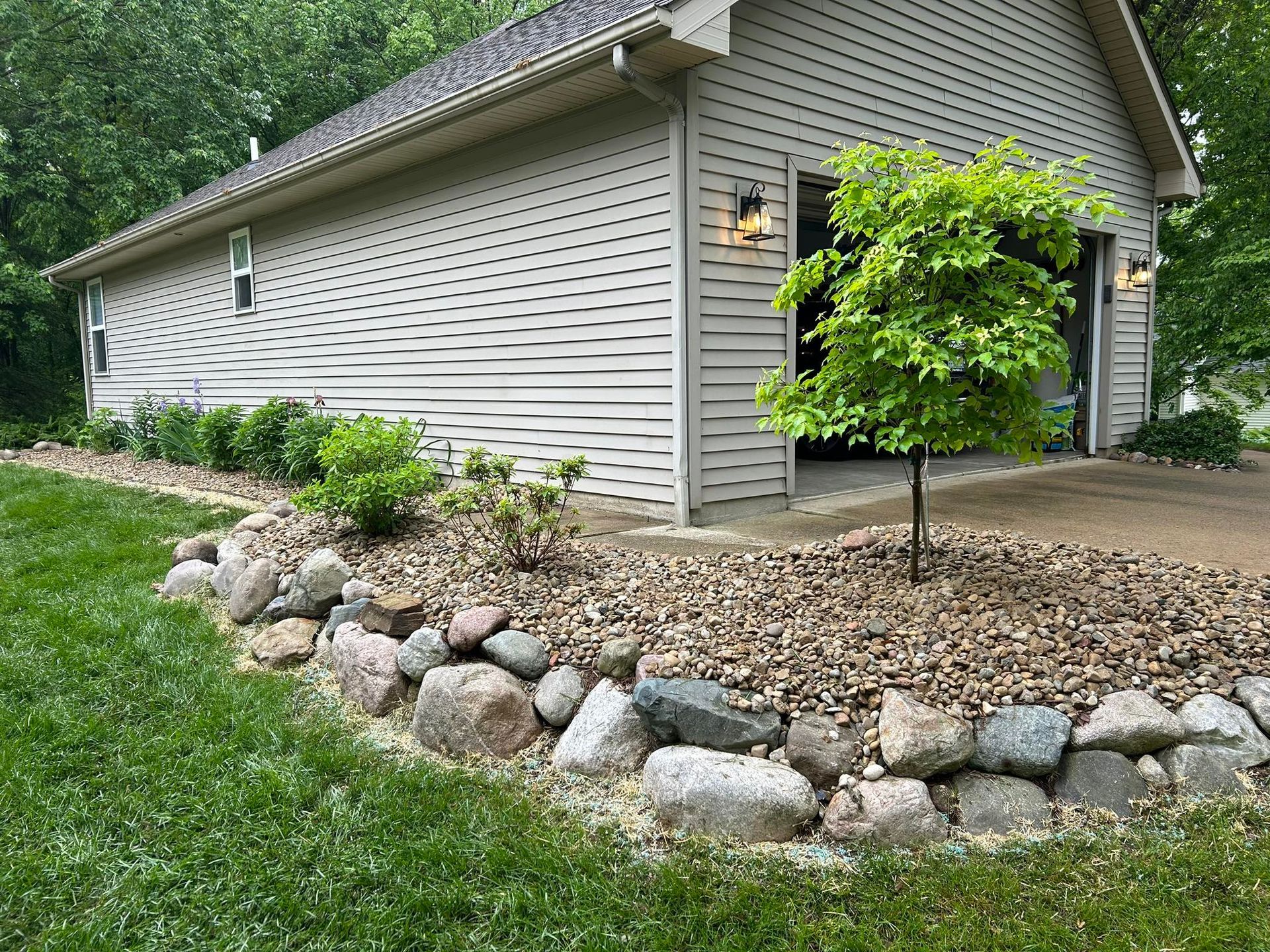 A light-colored vinyl-sided garage with a stone retaining wall and a small tree in a landscaped front yard.