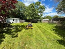 A yellow lawn mower sits in the center of a grassy backyard, with a white building and mature trees in the background.