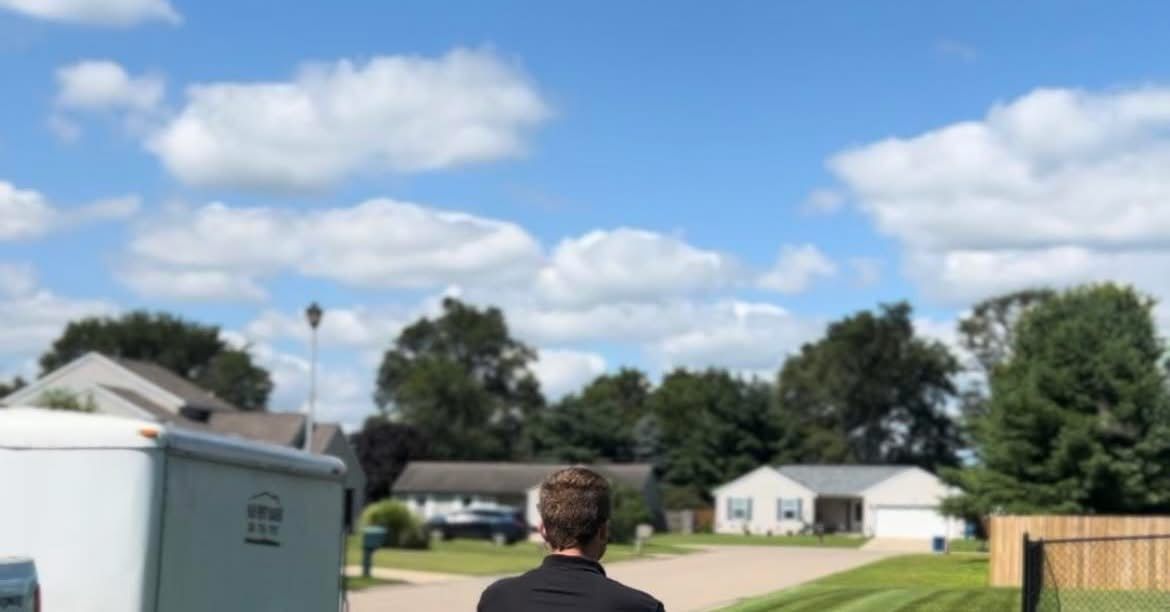 A person in a black shirt stands in a suburban street on a sunny, cloudy day, facing toward houses and trees.